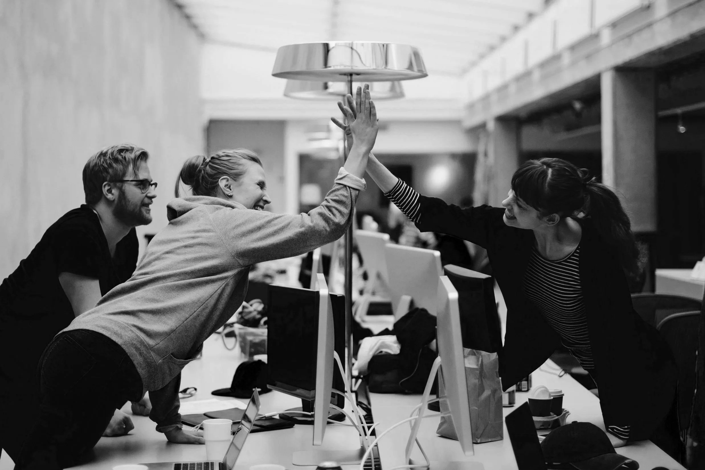 Two women and one man in an office giving each other a high five, with computer screens and desks around them.