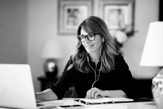 Woman with glasses smiling during a video call, sitting at a desk with a laptop, wearing earphones.