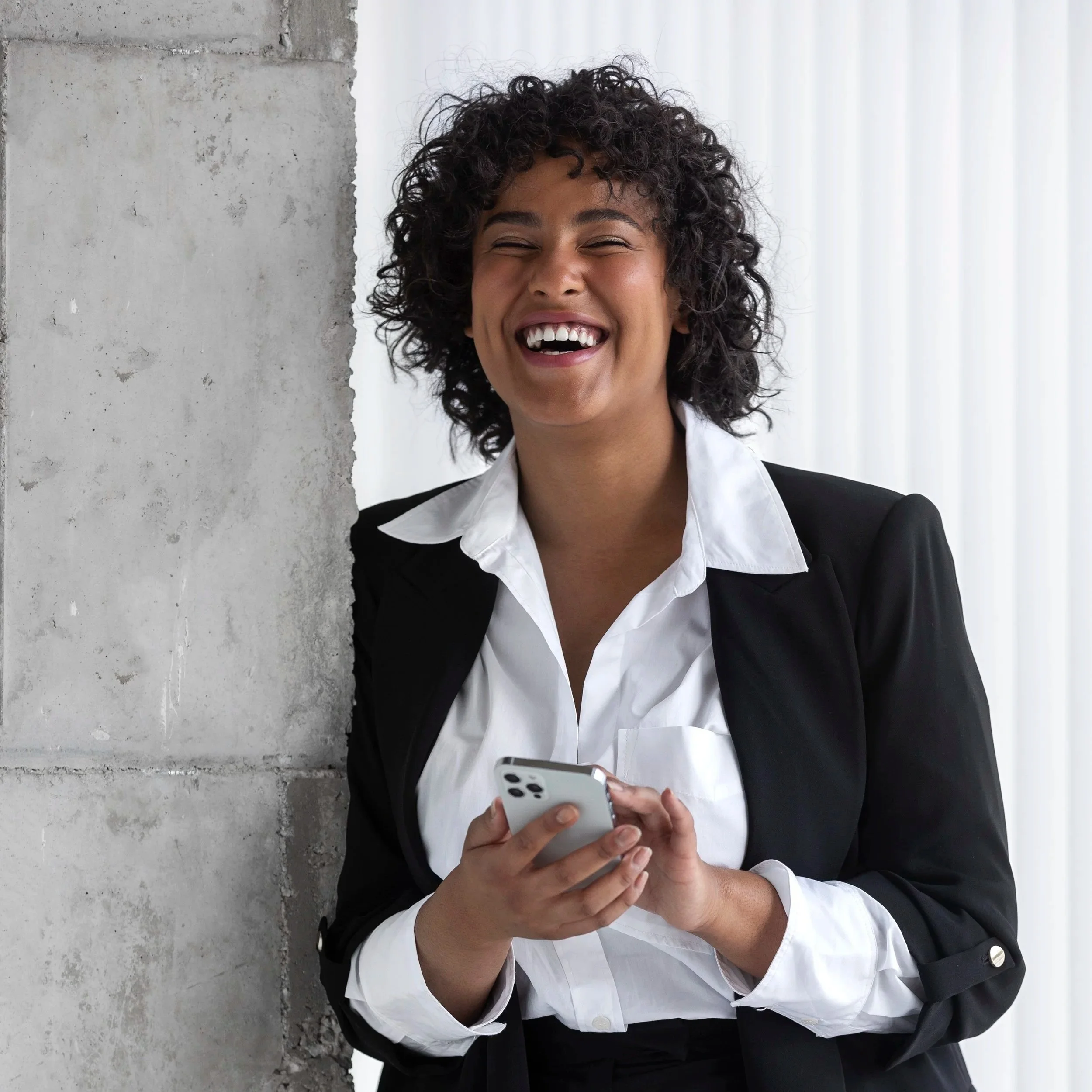 Woman with curly hair wearing a black blazer and white shirt looking at her phone and laughing.