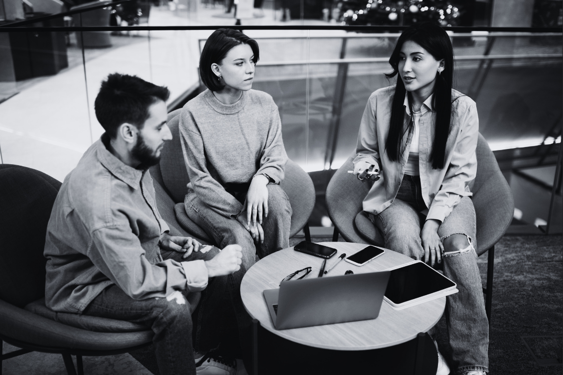 Three young adults sitting and talking around a small round table with laptops, phones, and a tablet in a modern indoor space.