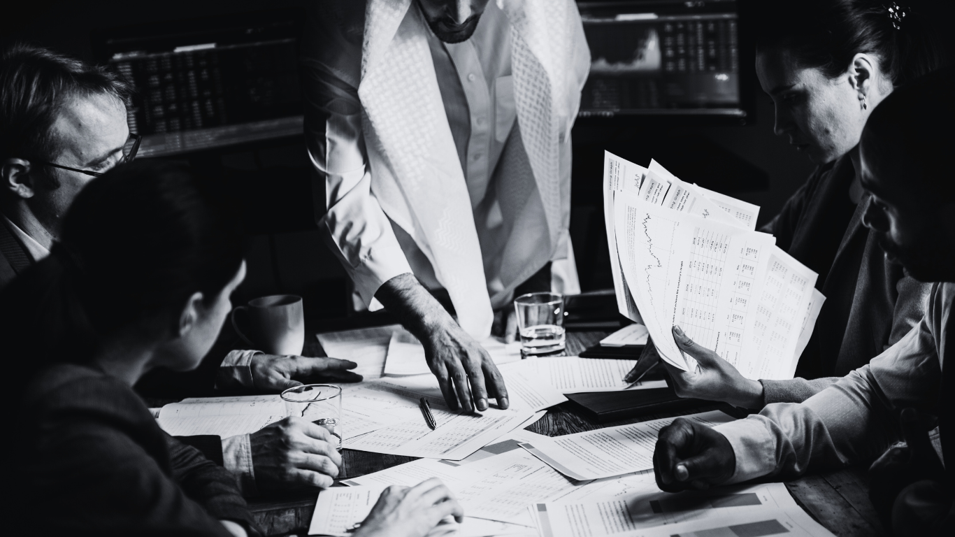 A group of five people gathered around a table, reviewing financial documents and charts in a business meeting. The scene is in black and white.