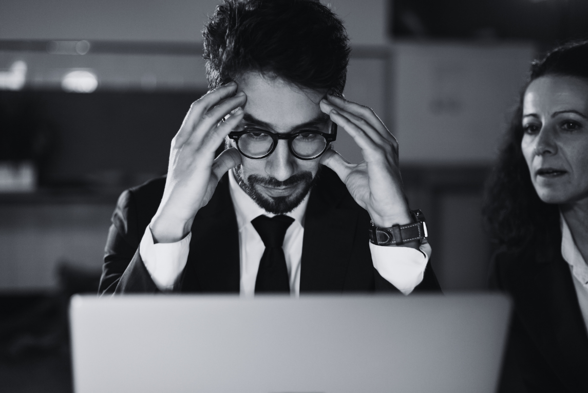 A man with glasses and a beard, dressed in a business suit and tie, sitting at a desk with a laptop, holding his head with both hands, appearing stressed or frustrated. A woman beside him is looking at the laptop, also appearing concerned, in a professional office setting.