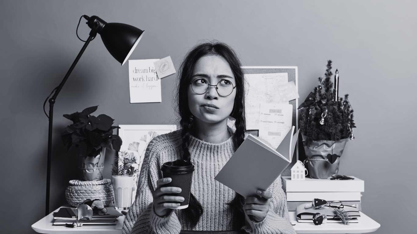 A woman with glasses, holding a coffee cup in one hand and an open book in the other, sitting at a cluttered desk with plants and papers. A lamp illuminates her workspace, and the background features a bulletin board with notes.