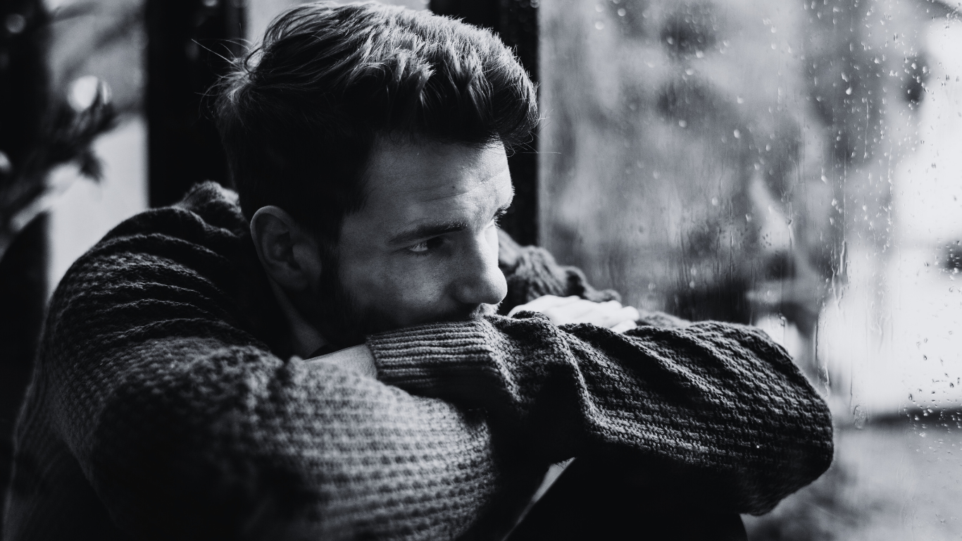A young man with dark hair and a beard leans on a window ledge, looking out with a somber expression. He is wearing a thick, textured sweater and appears thoughtful or melancholic in a black and white photo.