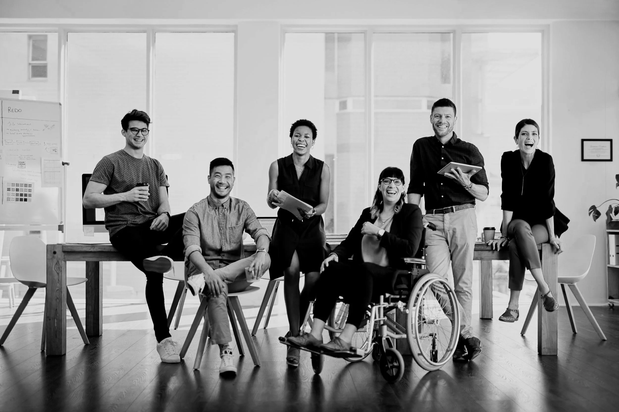 Group of six diverse coworkers smiling and laughing in an office, some seated and some standing, with large windows and office furniture in the background.
