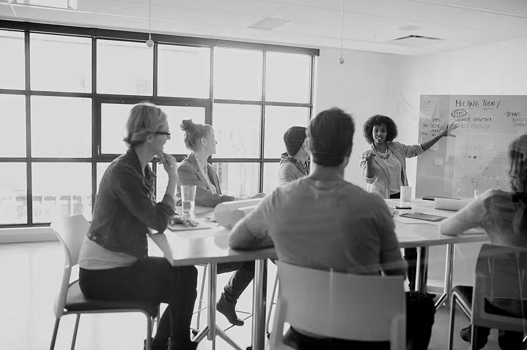 A diverse group of people in a meeting room, with a woman at the whiteboard giving a presentation to five other individuals seated around a table.