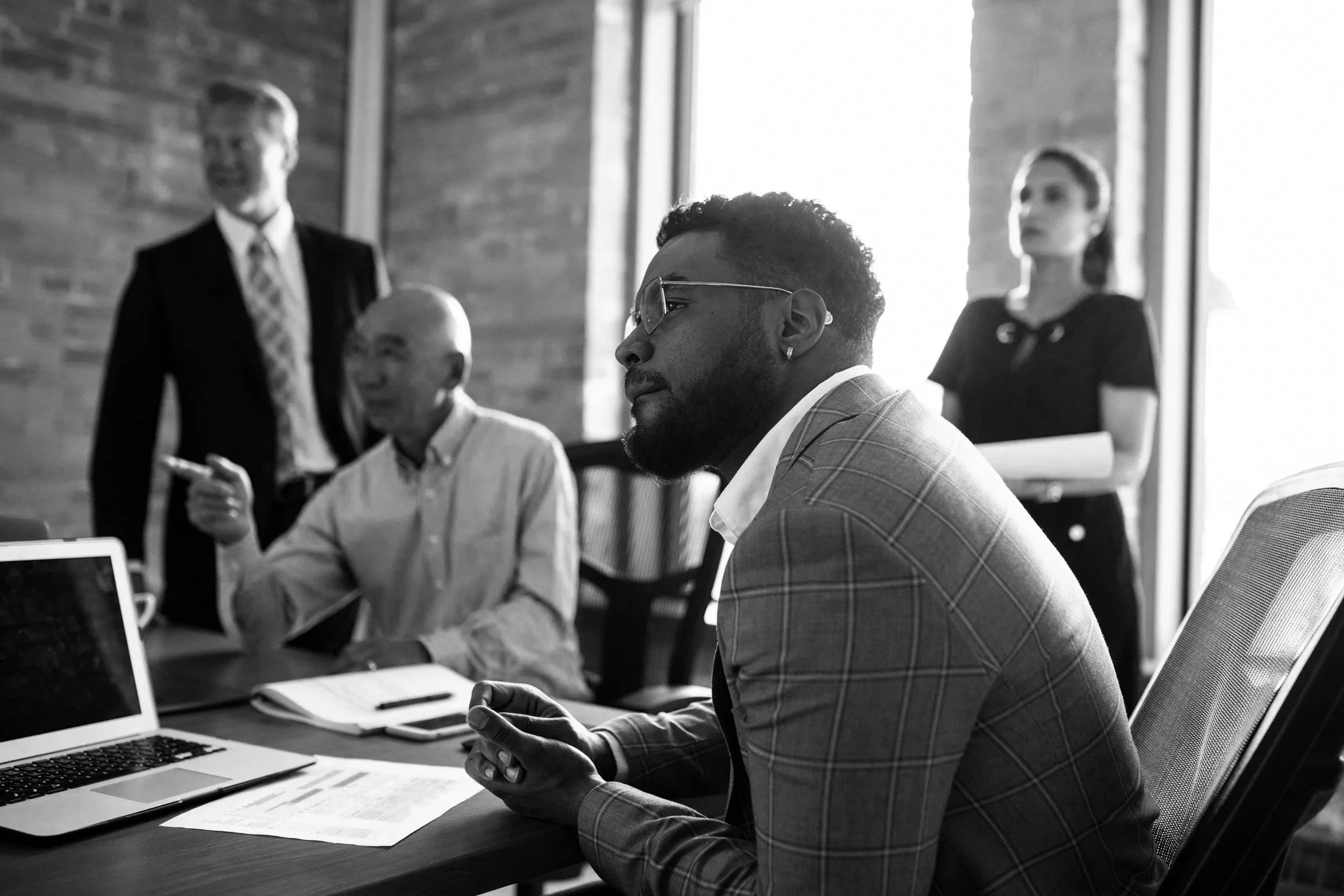 A business meeting in progress with a man in the foreground wearing glasses, a suit, and looking thoughtfully, surrounded by other professionals and a woman standing in the background.