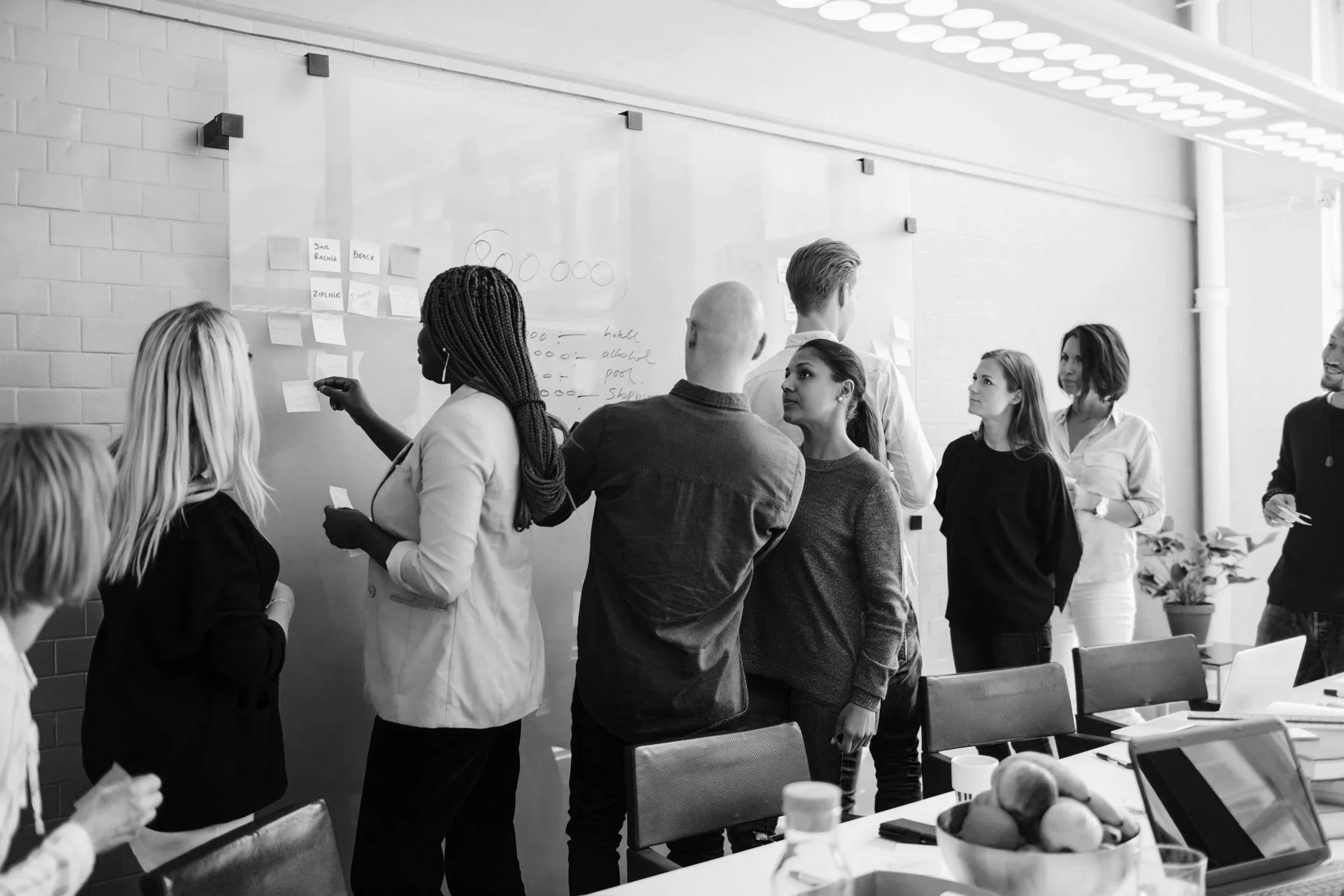 A diverse group of people collaborating in a business meeting in a modern office, discussing ideas on a whiteboard with notes and diagrams.