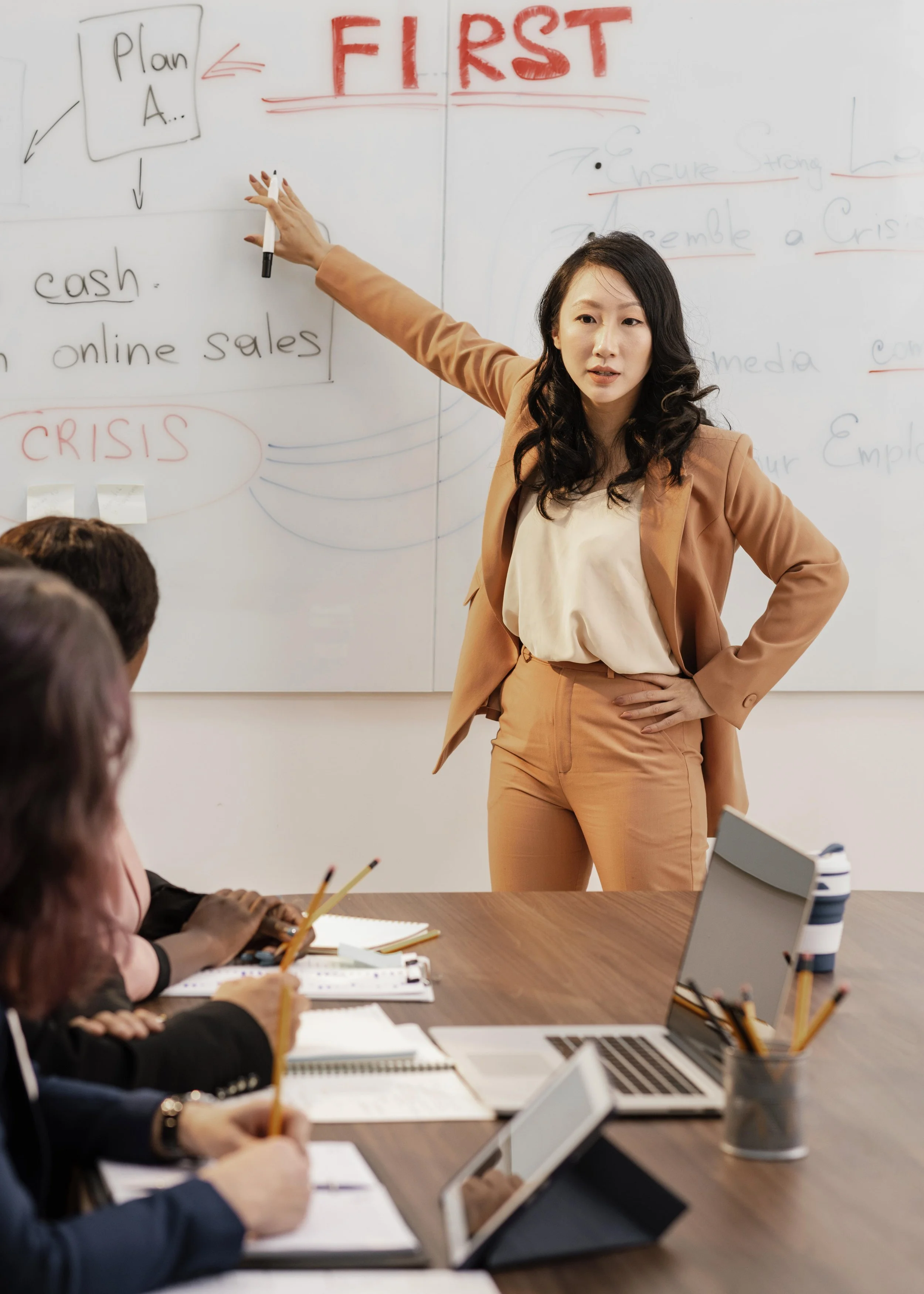 A woman in a tan suit is giving a presentation in front of a whiteboard, pointing at it with a marker. The whiteboard has handwritten notes including "Plan A...", "cash", "online sales", and the word "CRISIS" in red. Three people are seated at a conference table, taking notes and looking at the presenter with laptops, tablets, notebooks, and cups on the table.