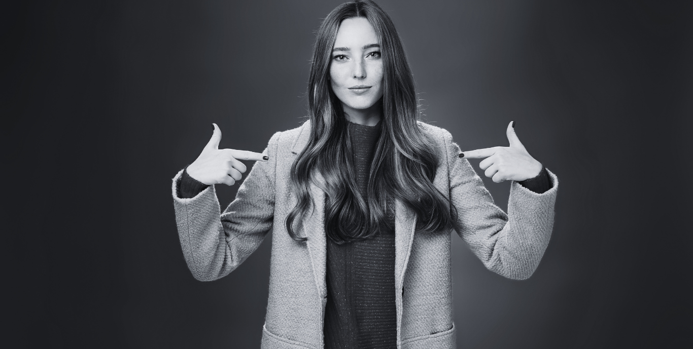 Black and white photo of a young woman with long wavy hair pointing at herself with both hands, wearing a coat and standing against a plain background.