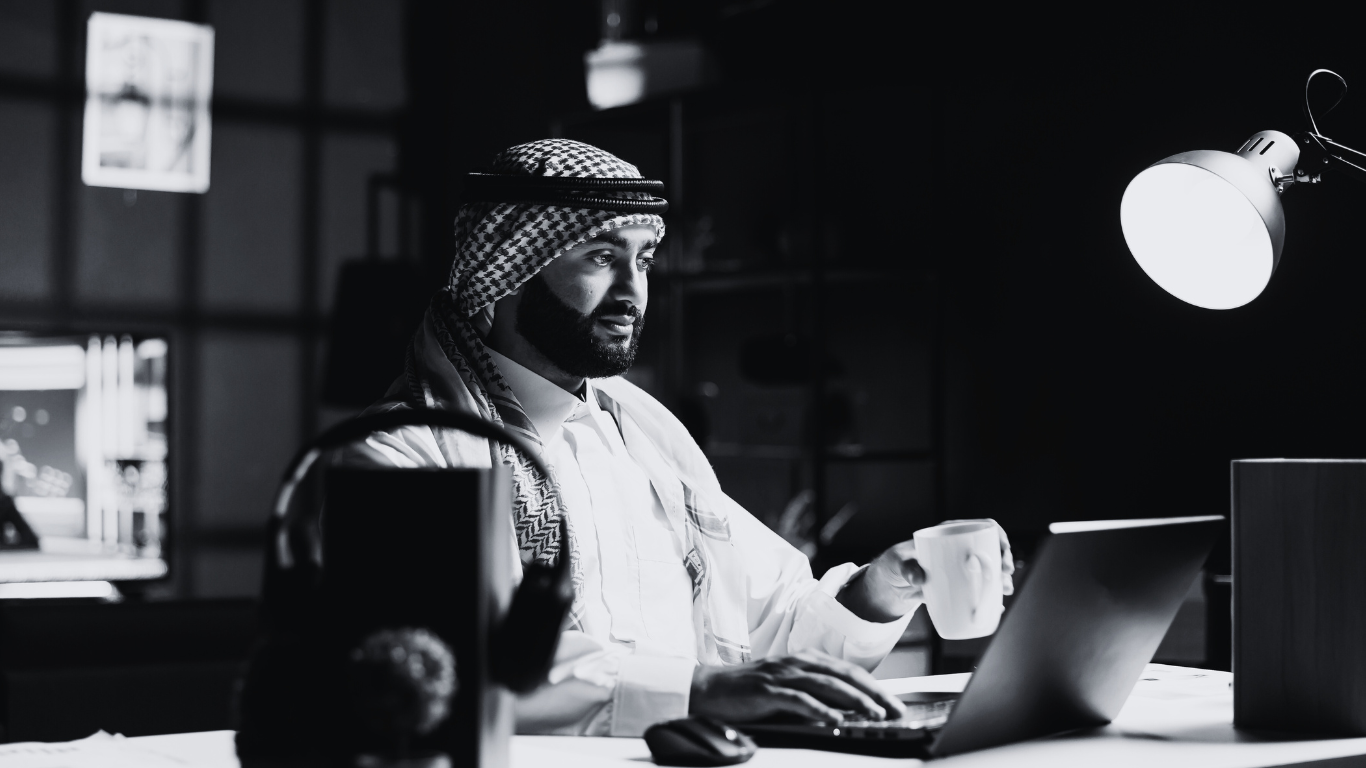 A man in traditional Middle Eastern attire sitting at a desk in a modern office, working on a laptop while holding a coffee mug, illuminated by a desk lamp.