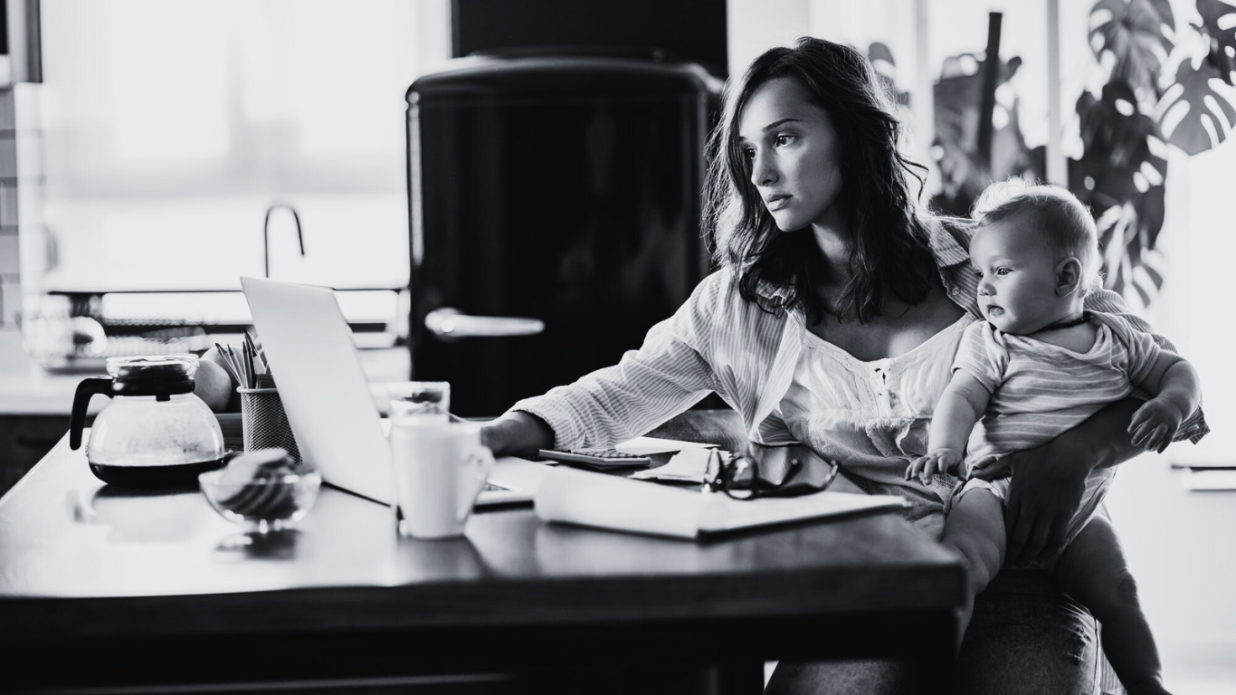 A woman sitting at a desk with a laptop, holding a baby on her lap, in a kitchen with various items on the table, in black and white.