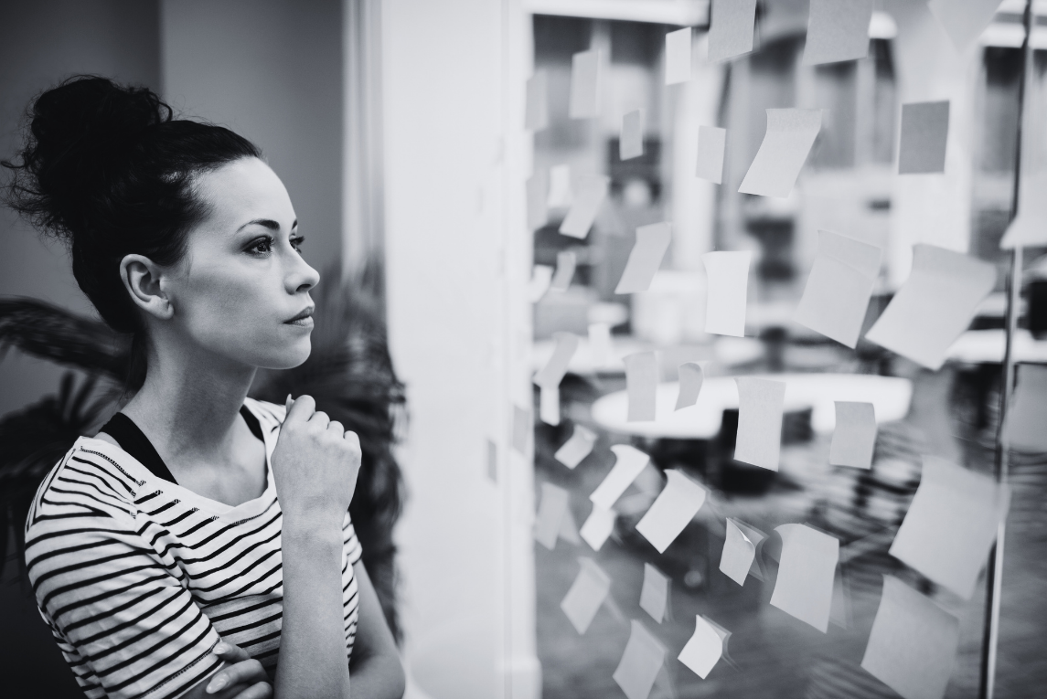 A woman in a striped shirt looking thoughtfully at a glass wall covered with scattered sticky notes in a modern office setting.