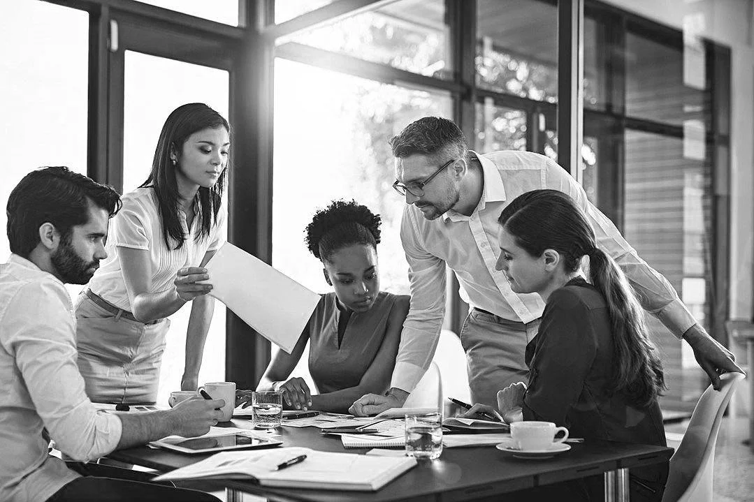 A group of six diverse professionals in a business meeting discussing around a table with documents, tablets, and glasses of water, inside a modern office with large windows.