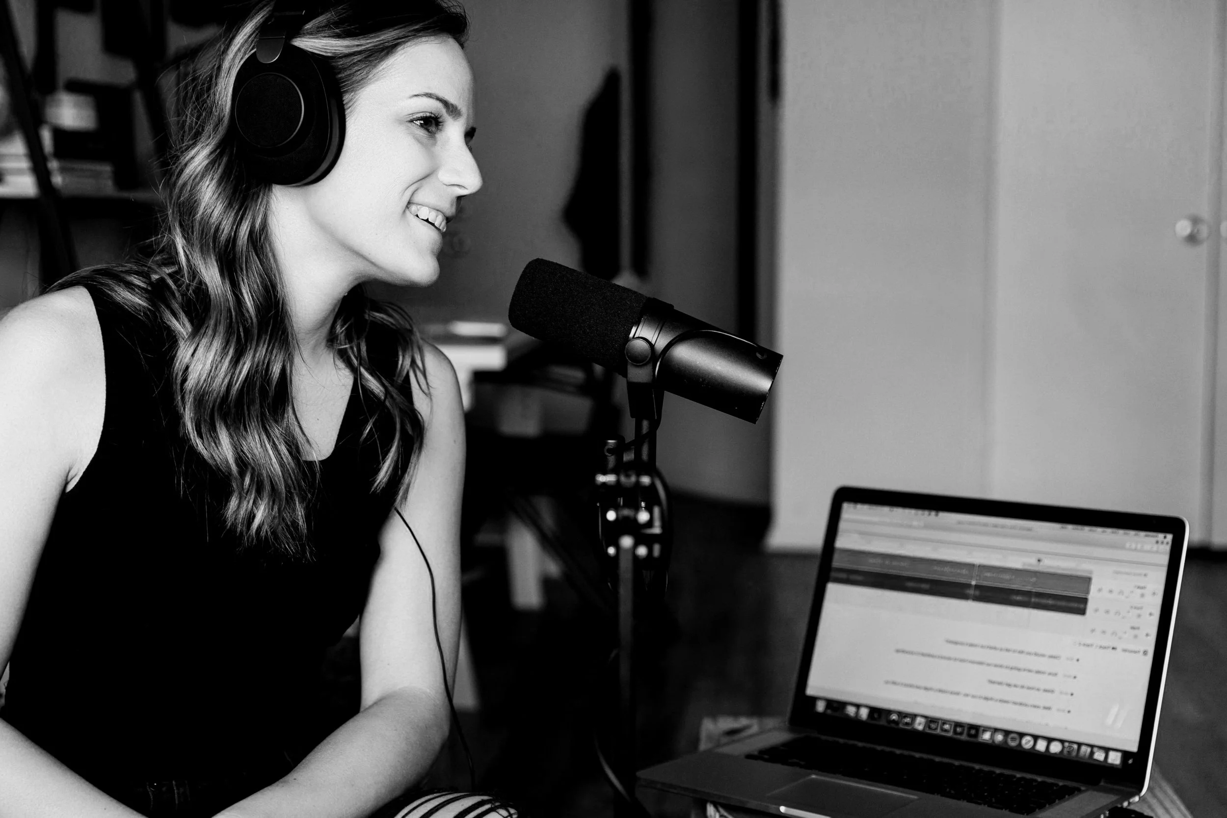 A woman wearing headphones speaks into a microphone while working on a laptop in a room.