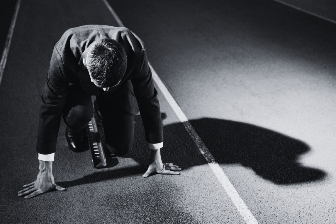 A person in a business suit in a starting position on a race track, ready to race, with shadows cast on the ground.