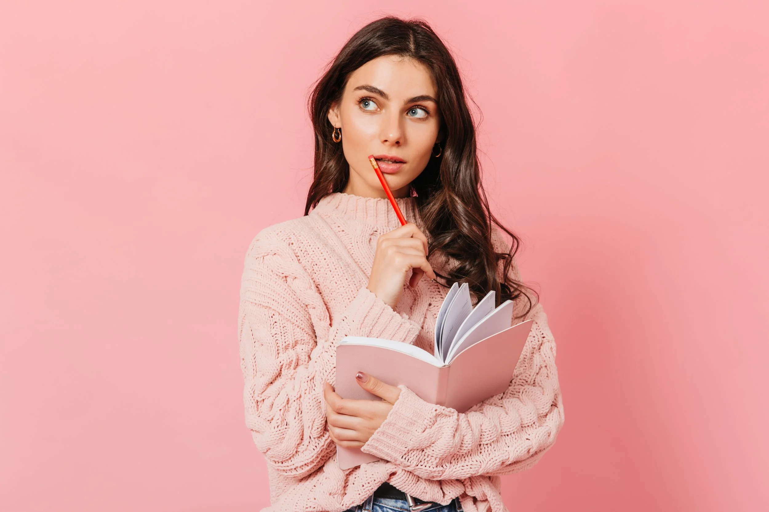 A young woman with long dark hair and blue eyes, wearing a pink knit sweater, holding a pink notebook, with a red pencil near her lips, thinking against a pink background.