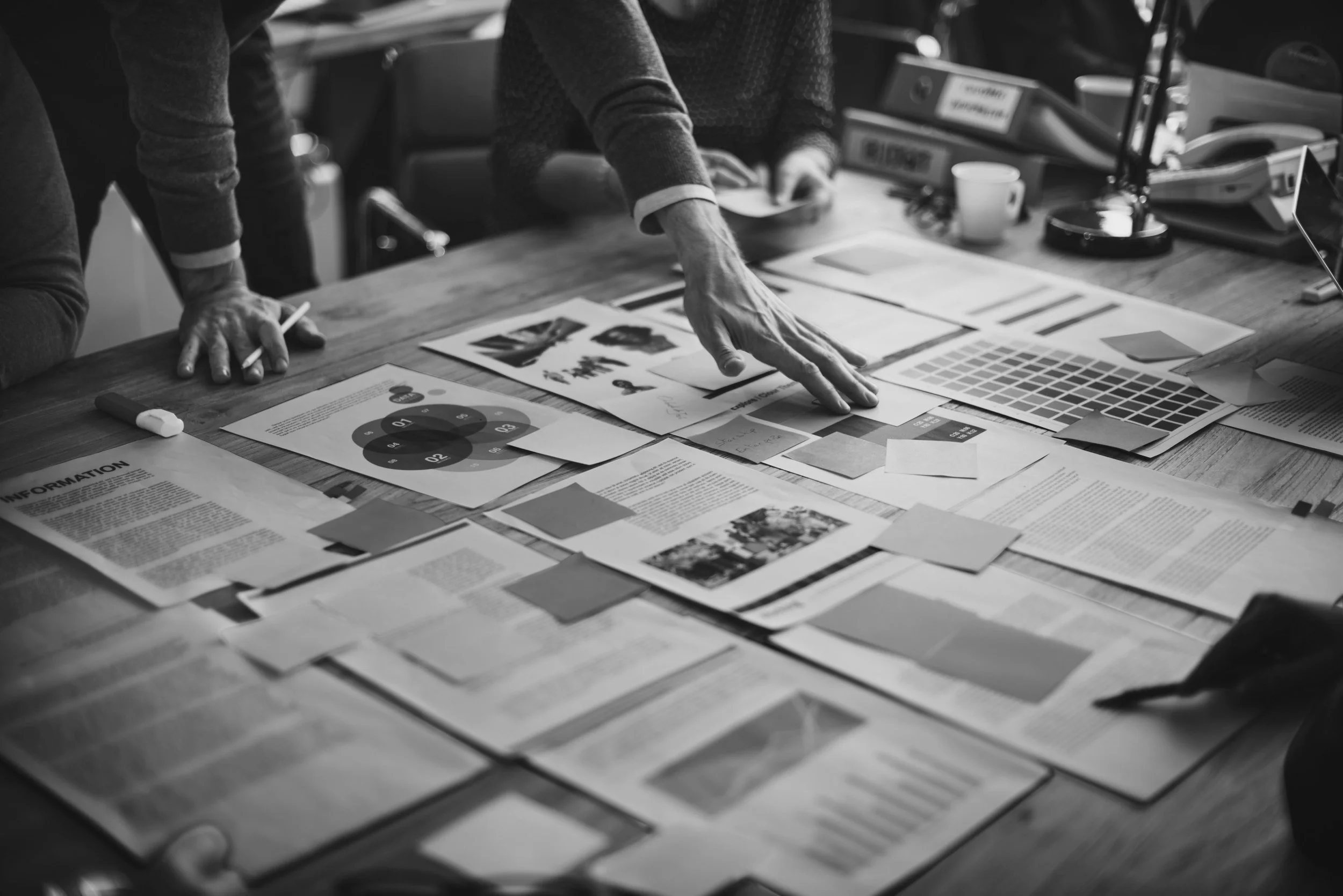 A black-and-white photo shows a group of people working at a cluttered desk with various documents, charts, and color swatches. One person's hand is pointing at a document, and another person's hand is on the table.