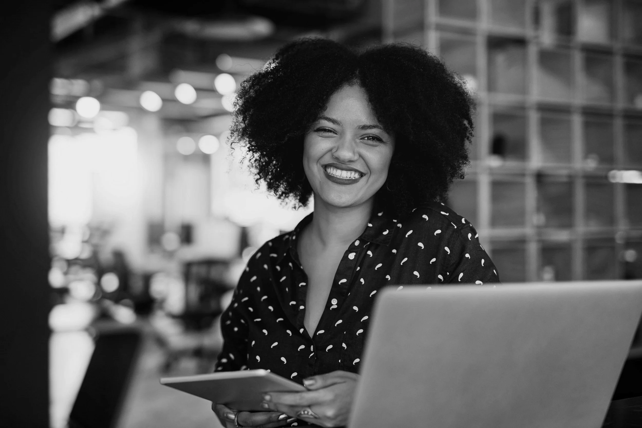 A smiling woman with curly hair, wearing a patterned shirt, sitting in a modern workspace with a laptop and a tablet.