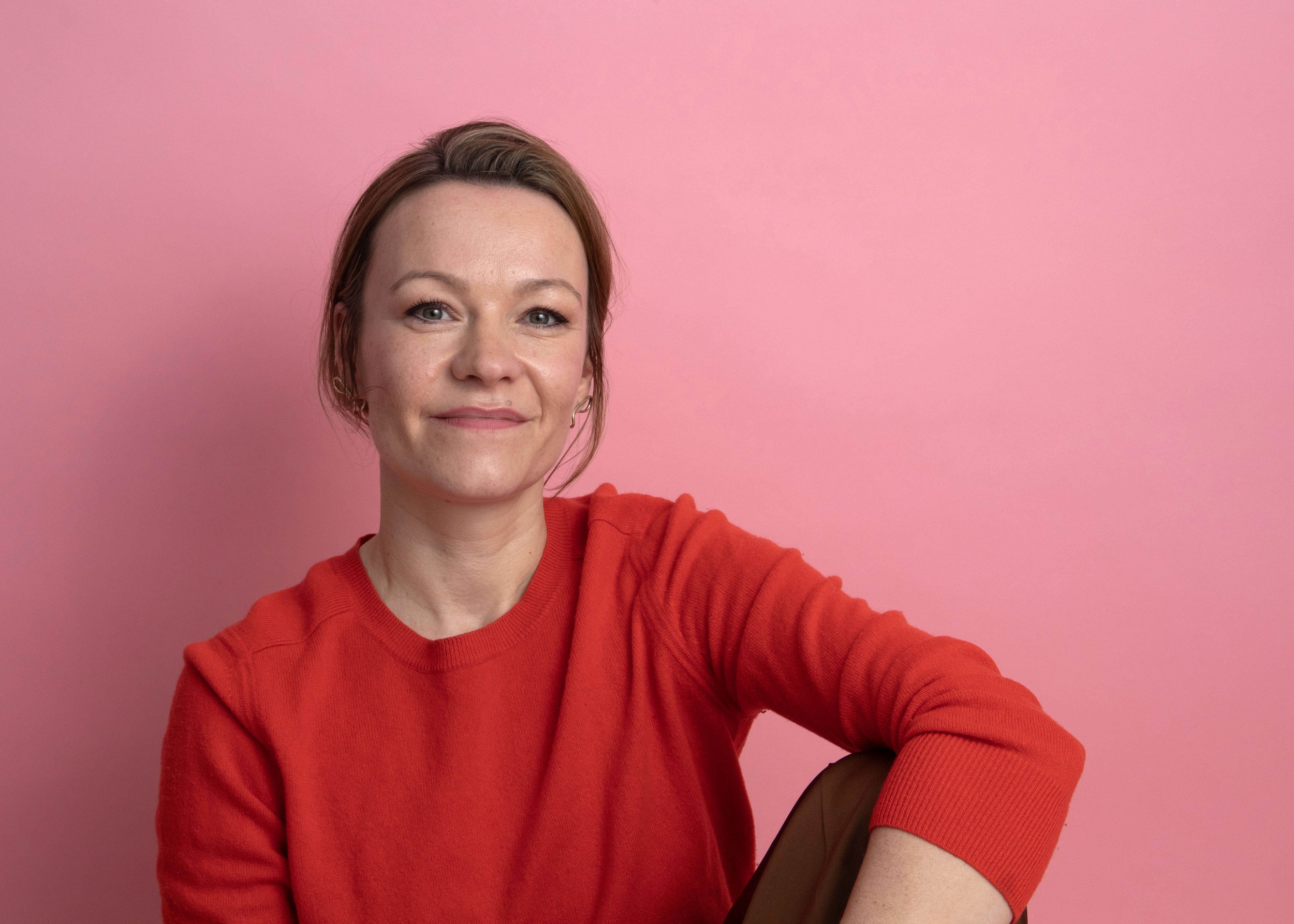 Kvinde med brunt hår, iført rød sweater, sidder mod en lyserød baggrund.