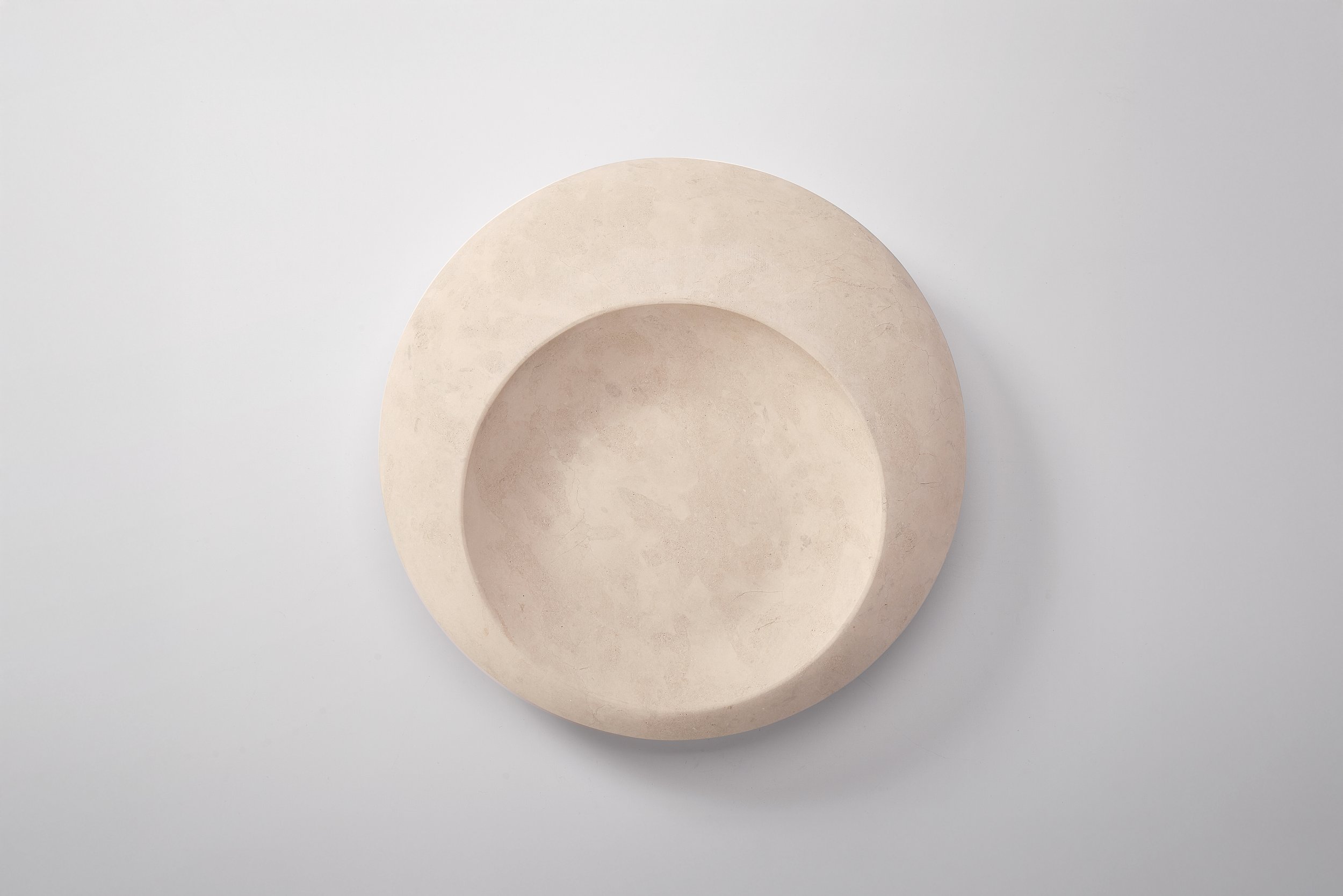 A minimalist, neutral-colored, concentric ceramic dish with a shallow bowl inside a larger, flat circular base on a plain light gray background.