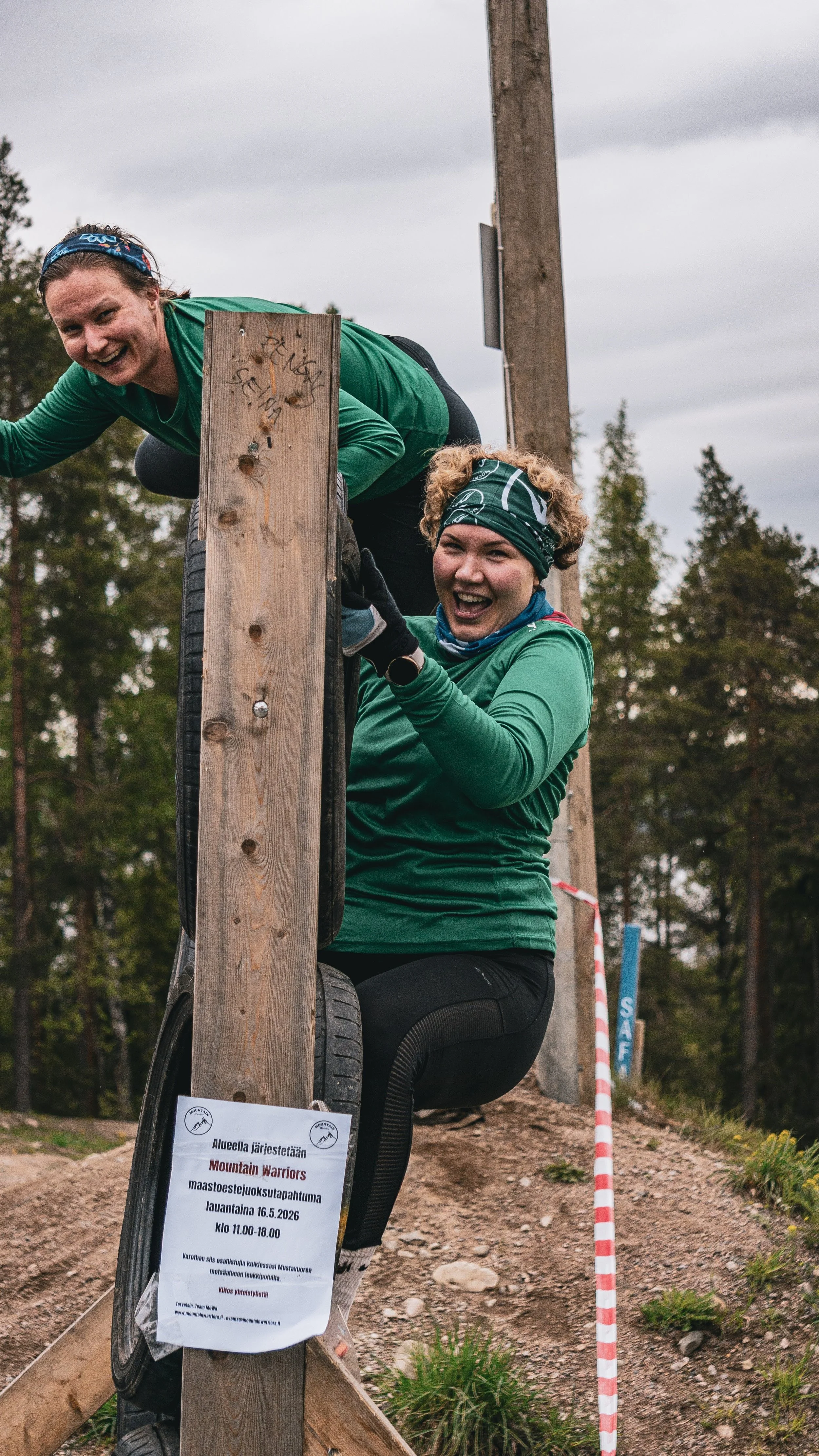 Kaksi naista kiipeää seinän yli yhteistyöllä. Molemmat ovat pukeutuneet samalaisiin tiimivaatteisiin ja bandanoihin. Taustalla puita ja Mustavuoren pilvinen taivas.