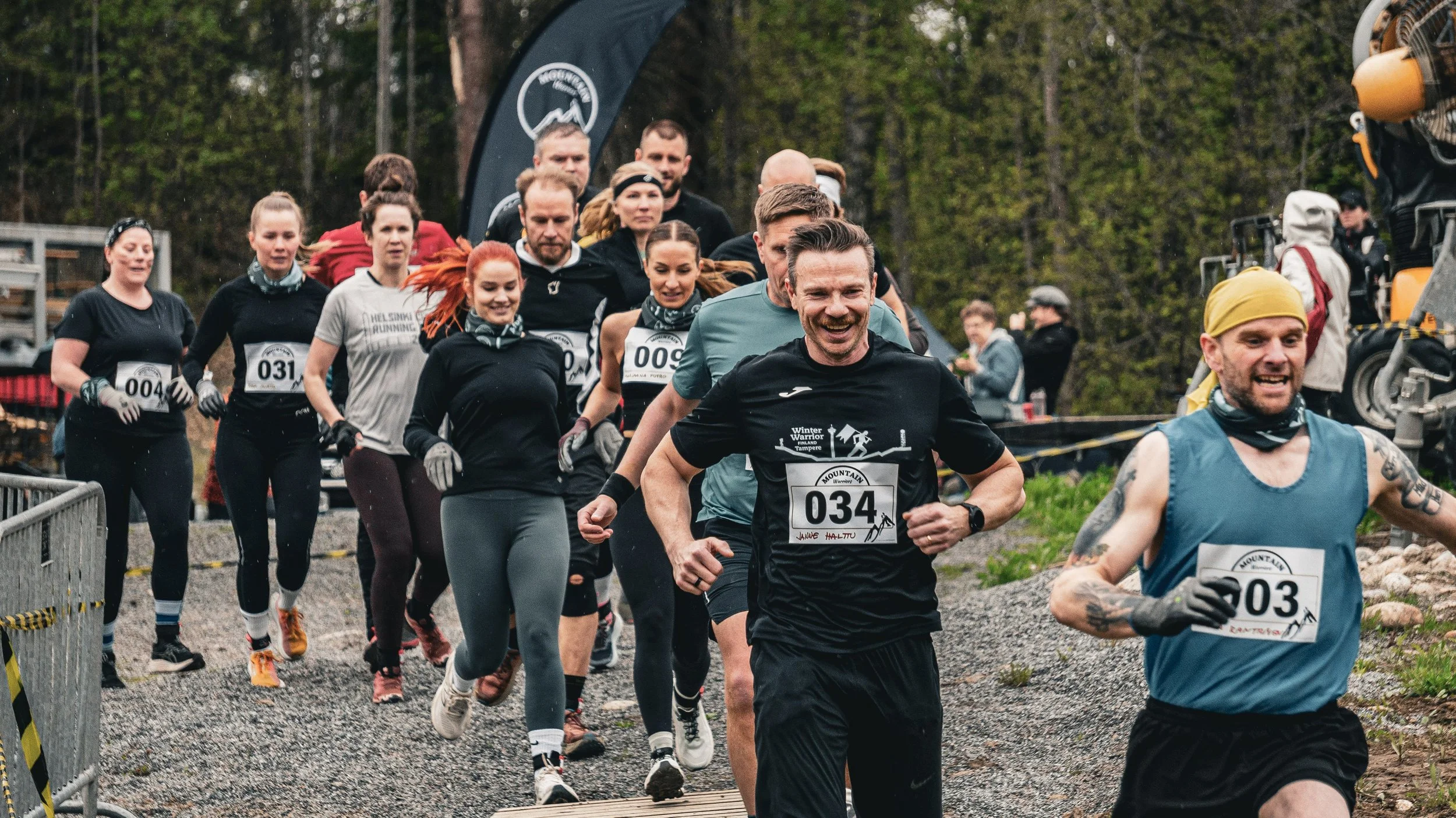 Juoksevia ihmisiä lähdössä kilpailemaan Mountain Warriors maastoestejuoksukilpailuun ulkona metsämaisemassa, iloisin ilmein.