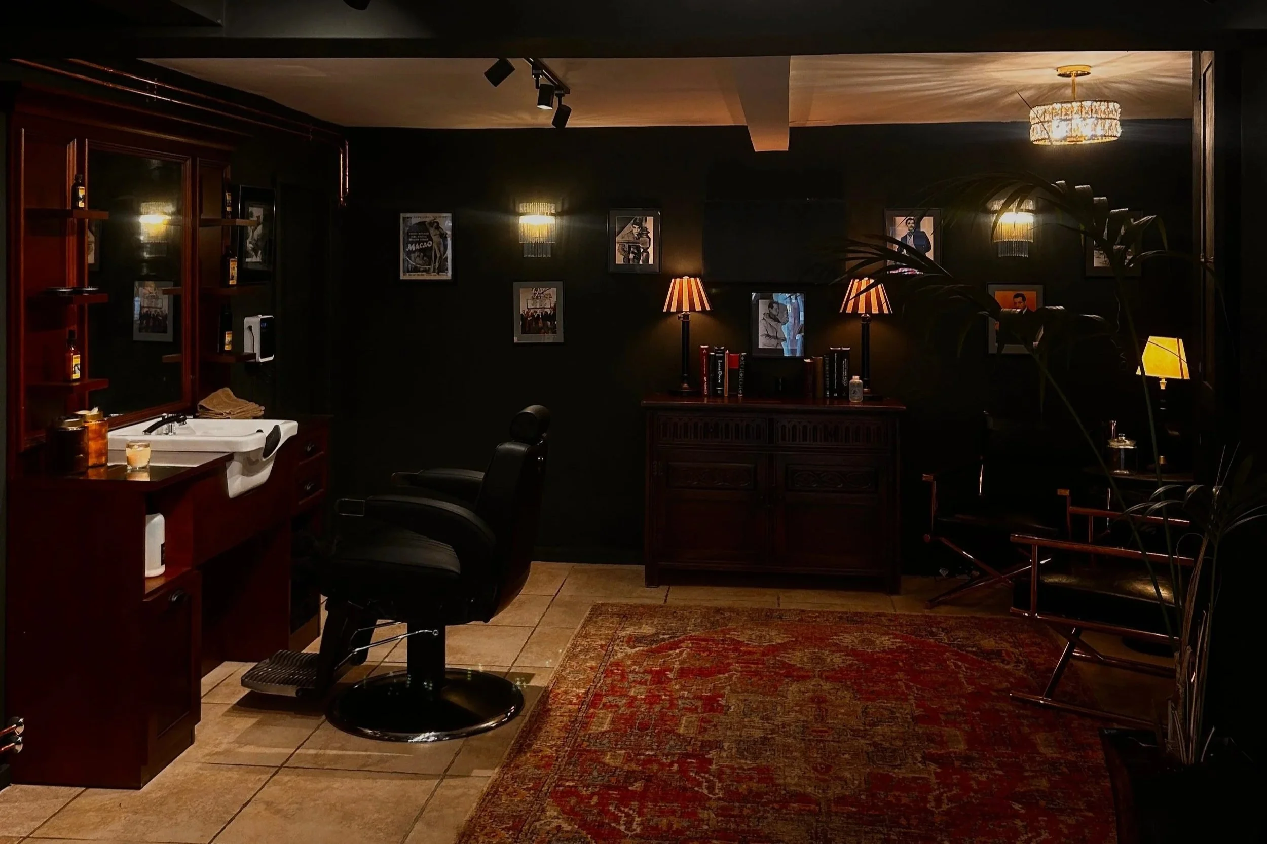 Dimly lit vintage barbershop or salon interior with black walls, wooden furniture, framed black-and-white photos, a leather barber chair, and warm lighting from lamps and chandeliers.