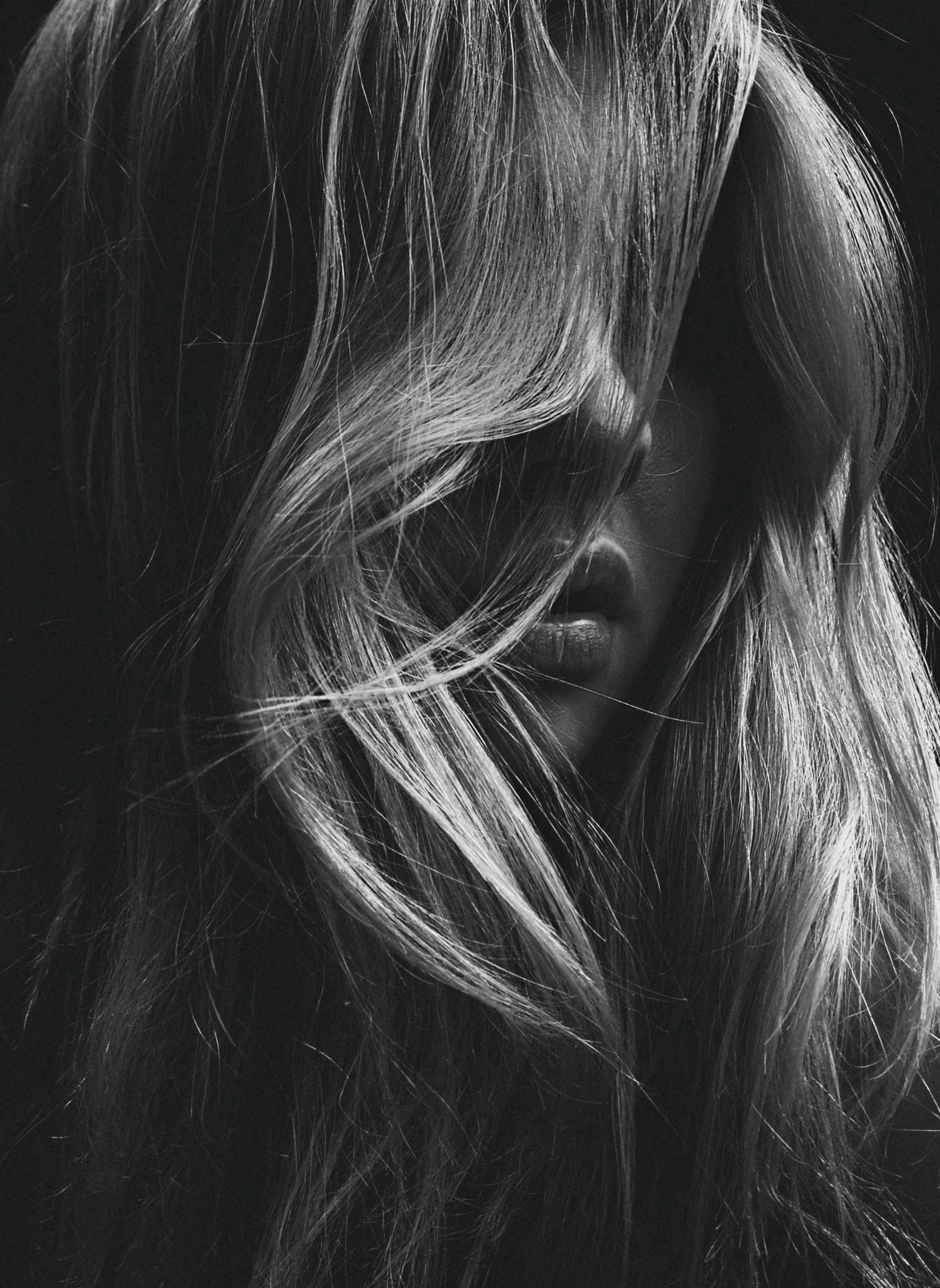 A black and white photograph of a woman with long, wavy hair partially covering her face, with a focus on her lips visible through the hair.