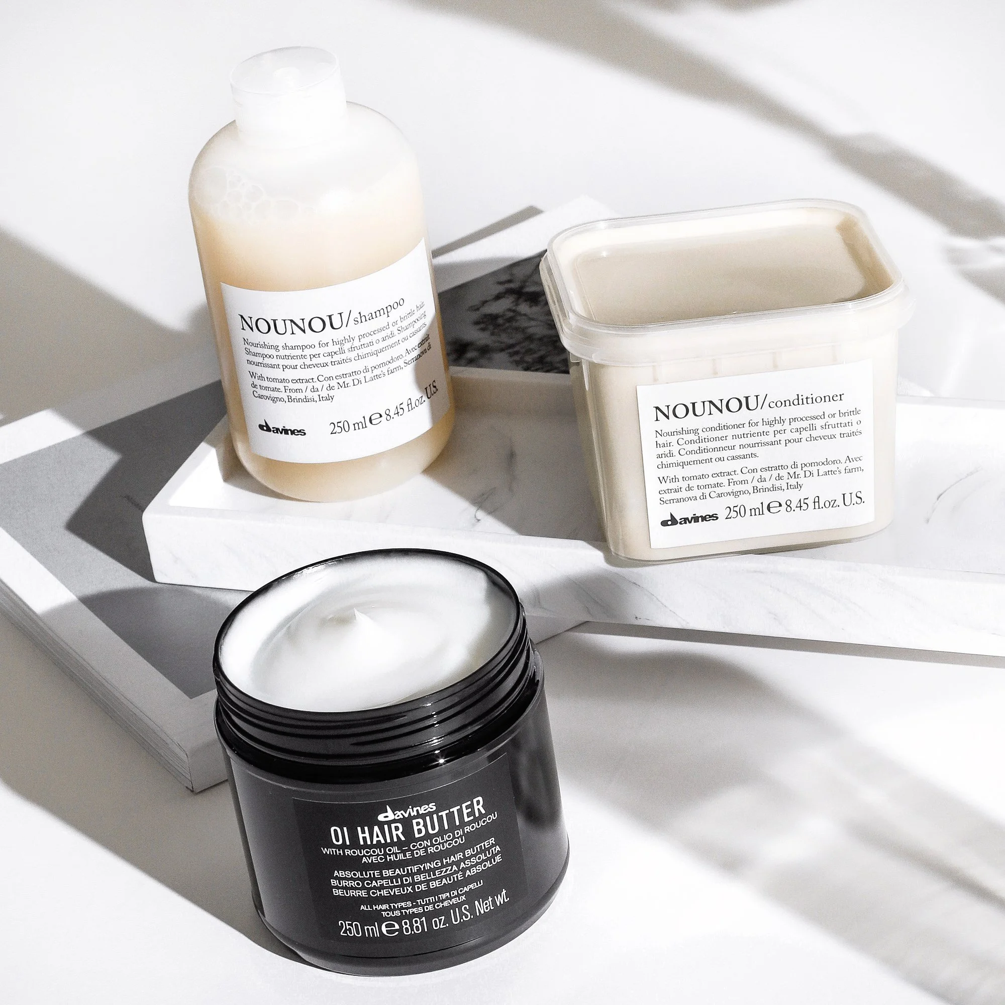Davines hair care products including NOUNOU shampoo, conditioner, and OI hair butter placed on white marble surface, with natural lighting and shadows.
