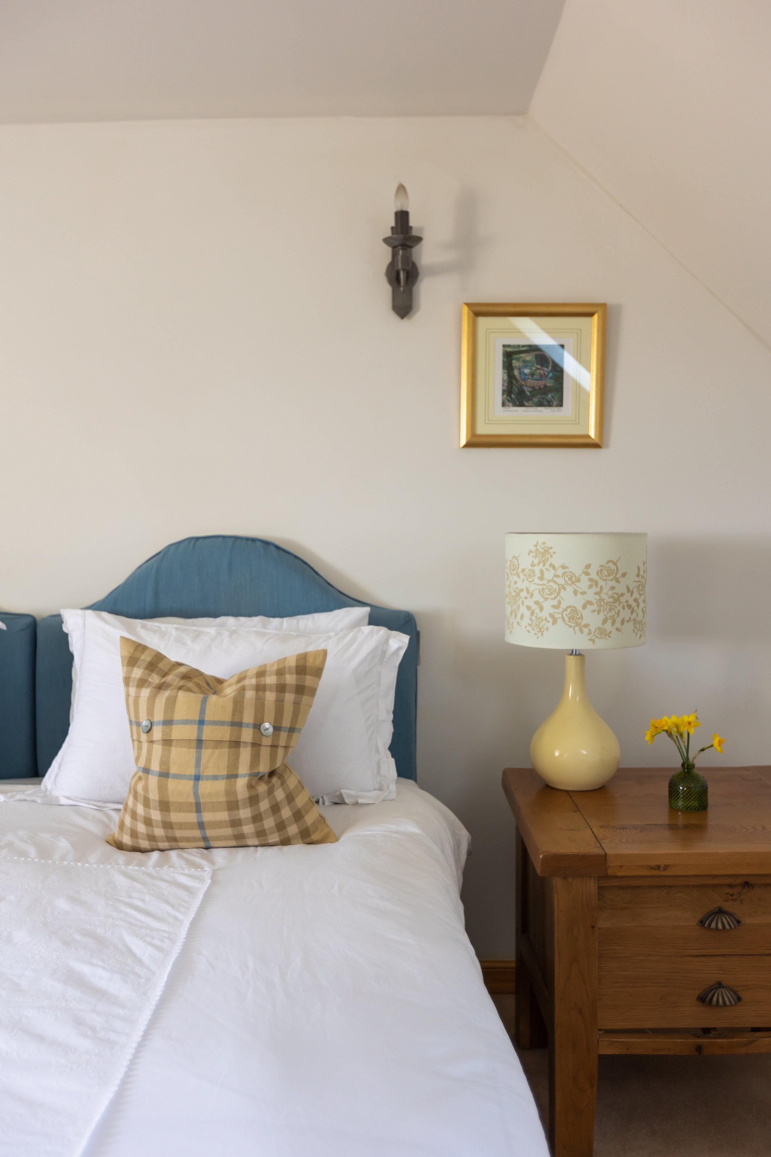 Bedroom with a blue upholstered headboard, white bedding, and a plaid cushion. A wooden nightstand with a cream lamp and small vase of yellow flowers is visible. A framed picture and wall sconce are above.