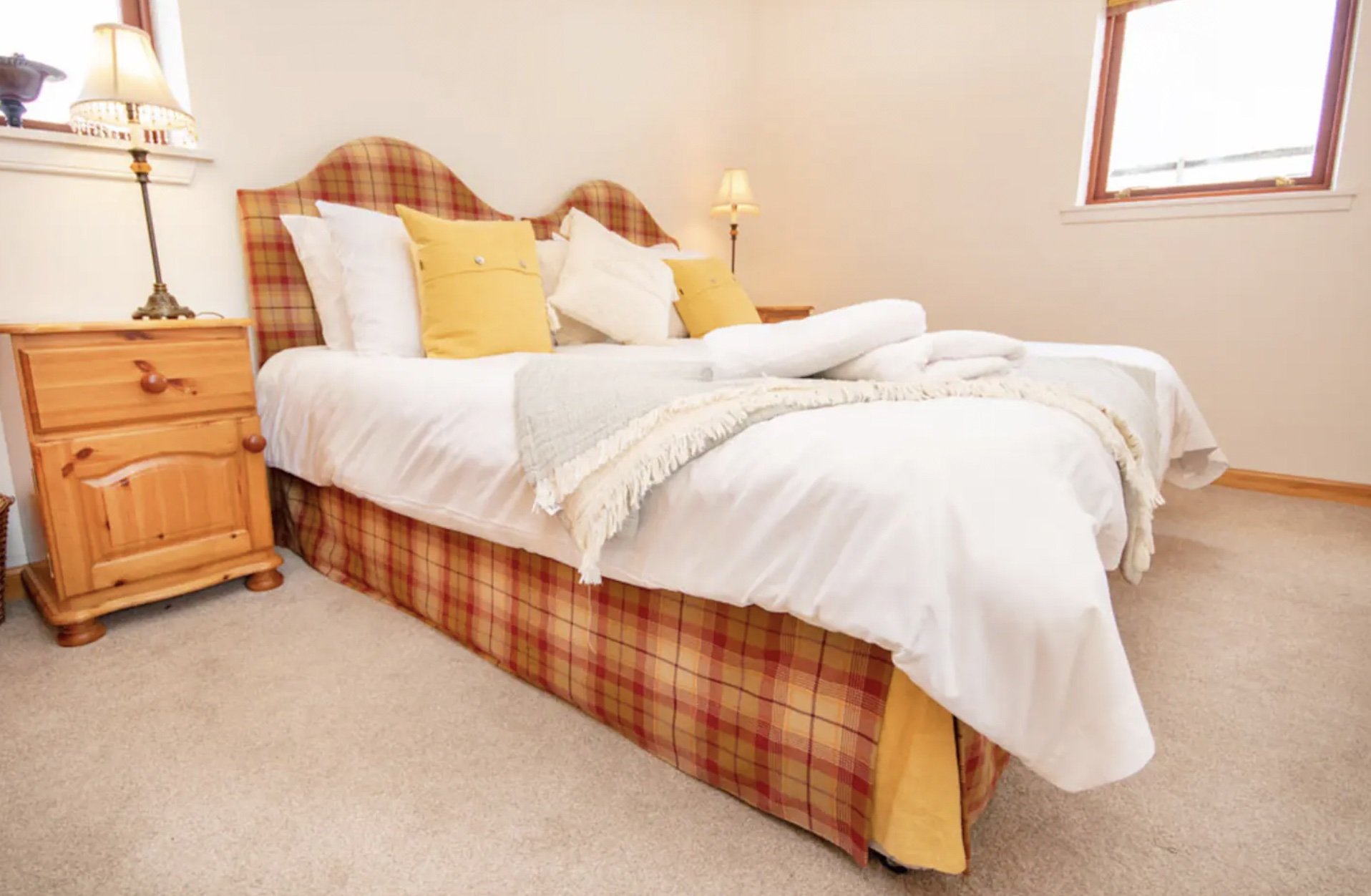 Cozy bedroom with a bed featuring plaid upholstery, white bedding, and yellow accent pillows. A wooden nightstand with a lamp is beside the bed. A window with natural light is above the headboard.