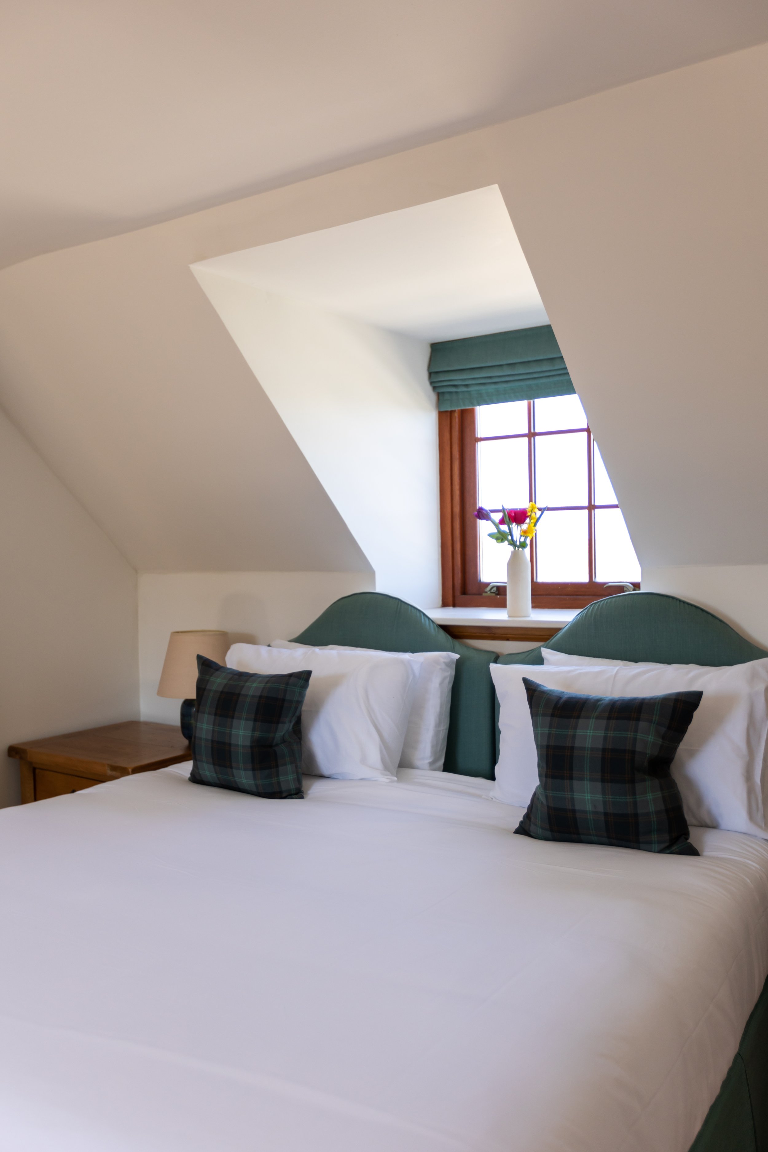 Cozy bedroom with white bedding, plaid pillows, and green upholstered headboard, featuring a wooden bedside table with a lamp, beneath a window with a vase of flowers.