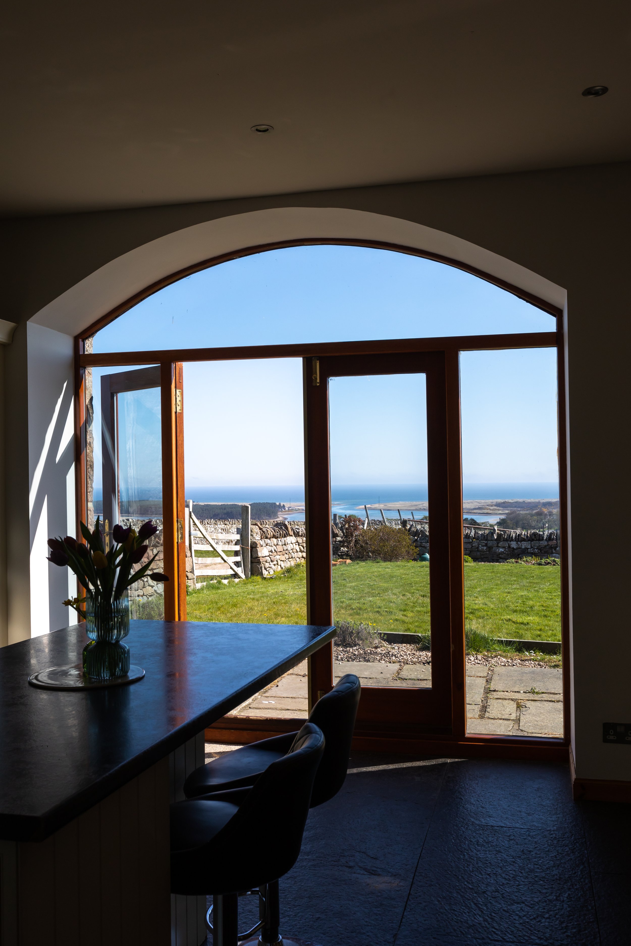 View from inside a room with an arched glass door opening to a garden and ocean view, featuring a countertop with a vase of flowers and stools.
