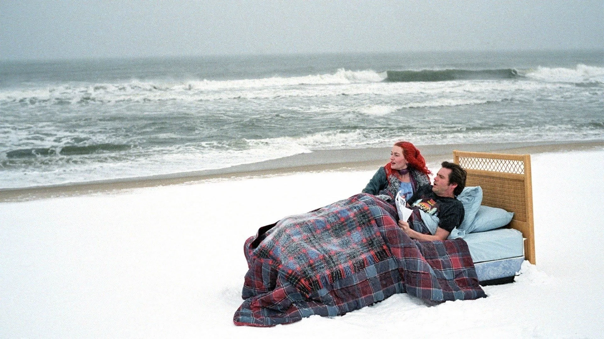 A couple lying in bed on a snowy beach with ocean waves in the background, the woman with red hair and the man reading a magazine.