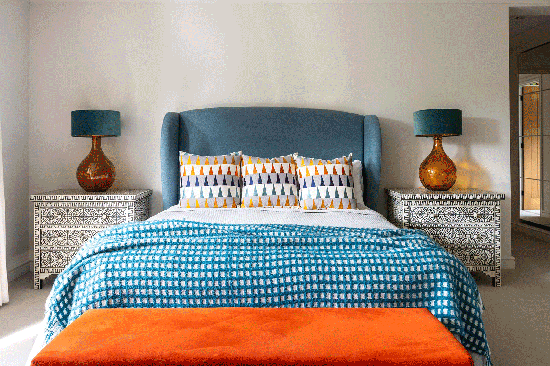 a giff of a light teal headboard withe three cushions with small orange details and glass orange bedside lights