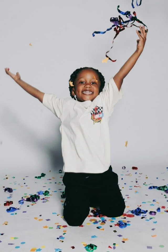 A smiling young girl with braided hair kneeling on the floor with arms raised, surrounded by colorful confetti and streamers, in front of a plain white background.