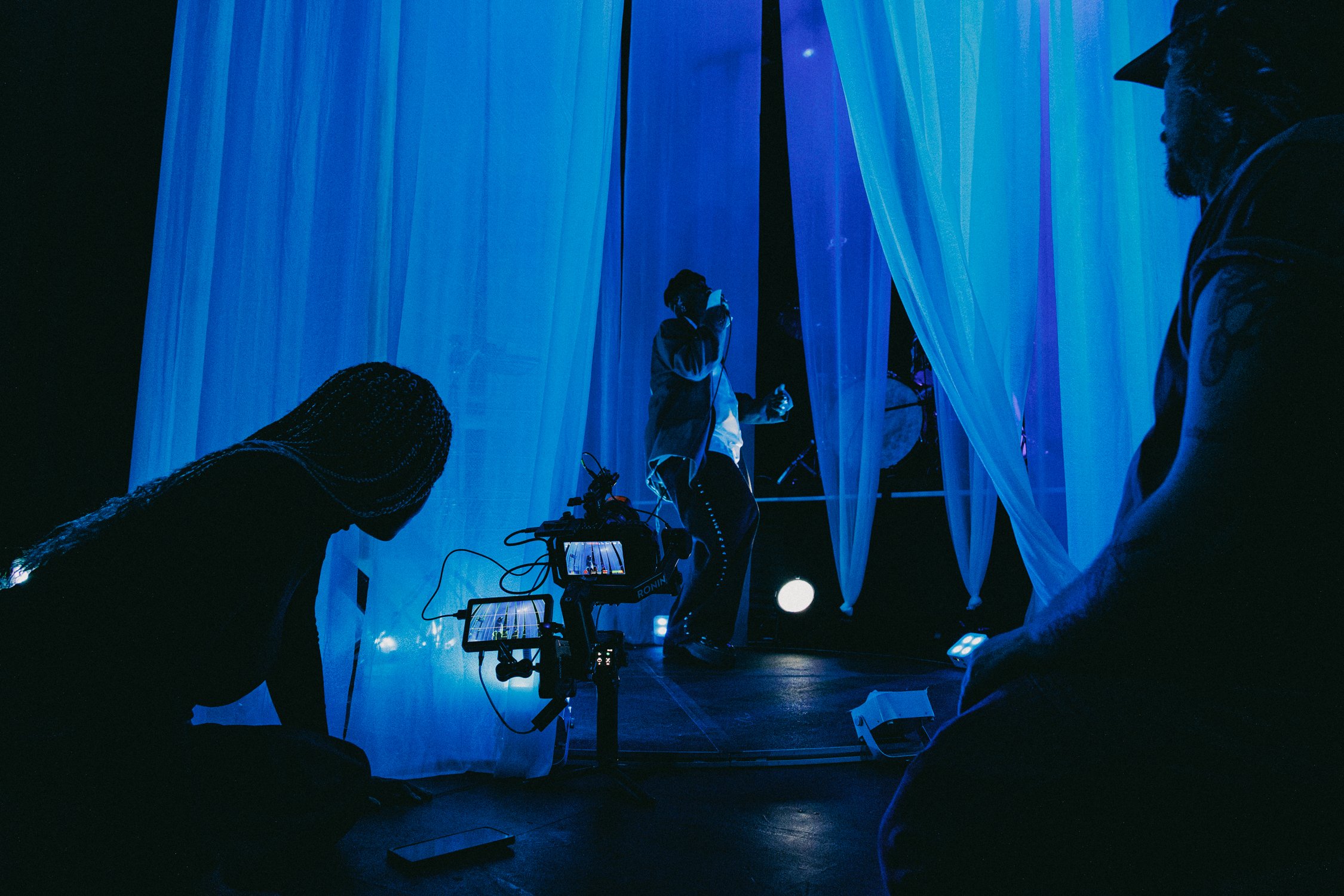 Filmmakers or photographers setting up a scene with blue lighting and sheer curtains, with a person filming on a camera and others coordinating behind the scenes.