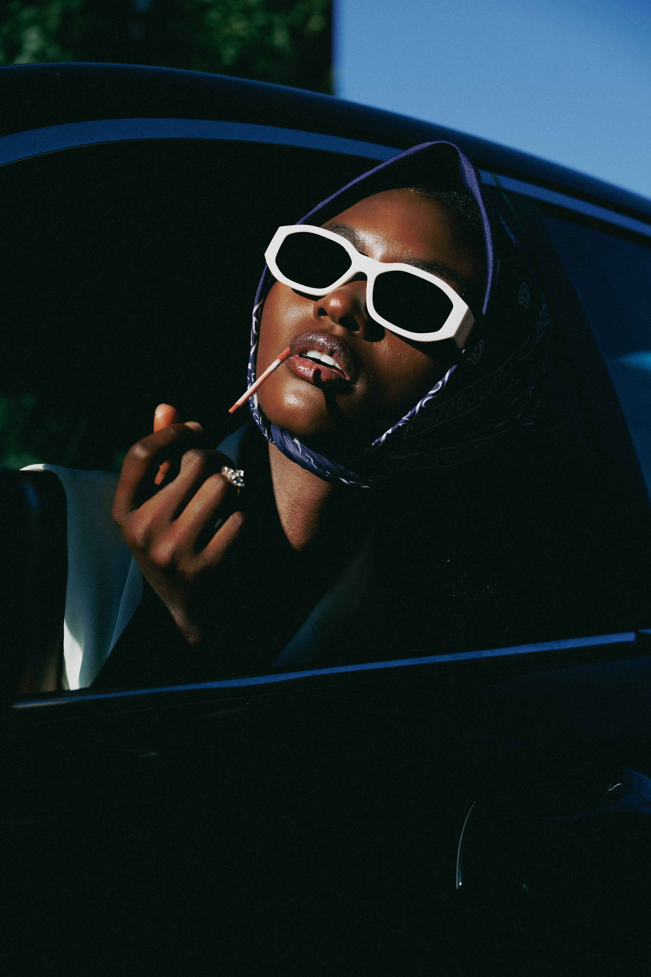 A person with dark skin and full lips wearing exaggerated white sunglasses, a dark bandana, and a jacket, sitting inside a car with the window down. The person is holding a lollipop and appears to be posing for the photo, with sunlight casting shadows on their face.