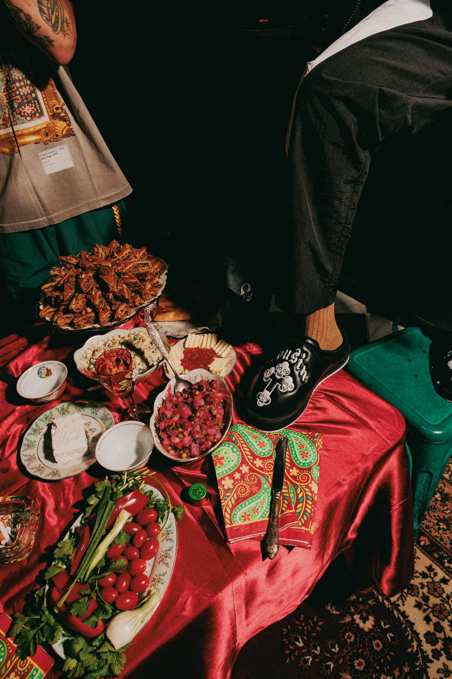 A table covered with a red satin cloth, holding various dishes including cherry tomatoes, herbs, salads, cheese, and dessert, with a person standing nearby in dark clothing