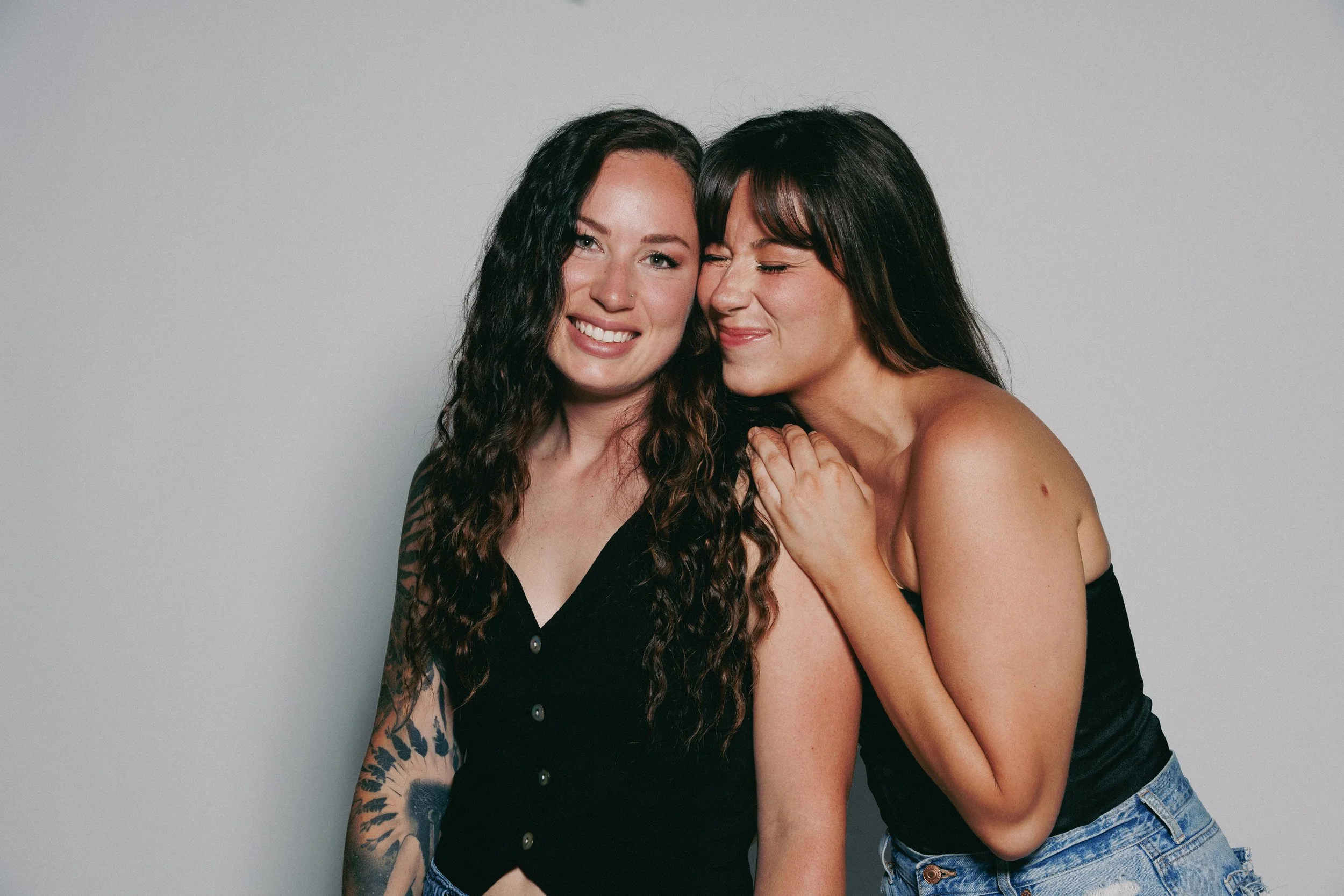 Two women smiling and hugging against a plain gray background.