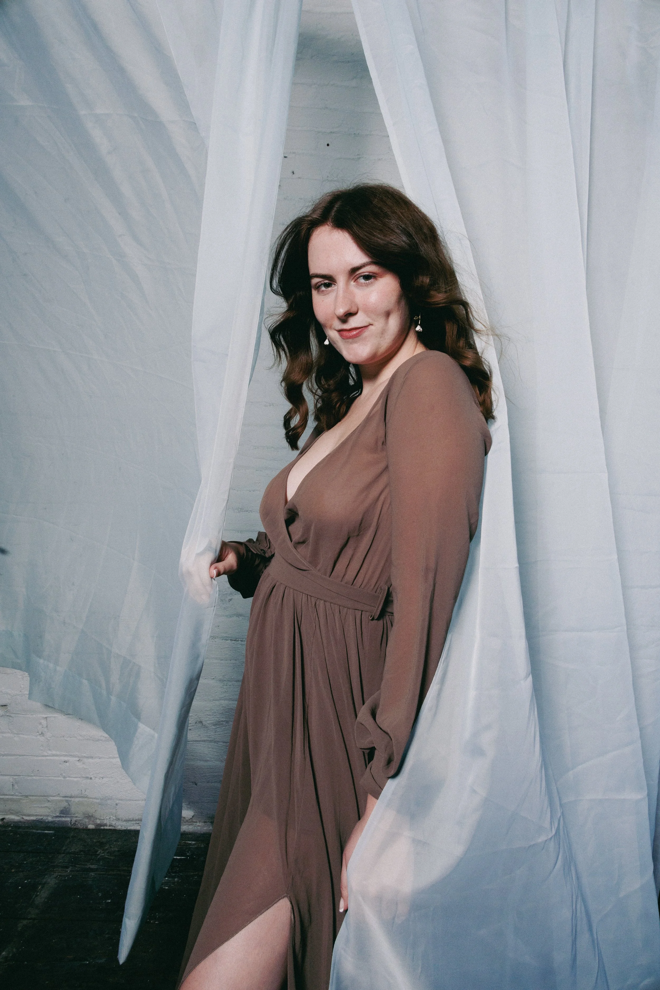 A woman with dark brown, wavy hair wearing a brown dress standing behind sheer white curtains with a white brick wall in the background.