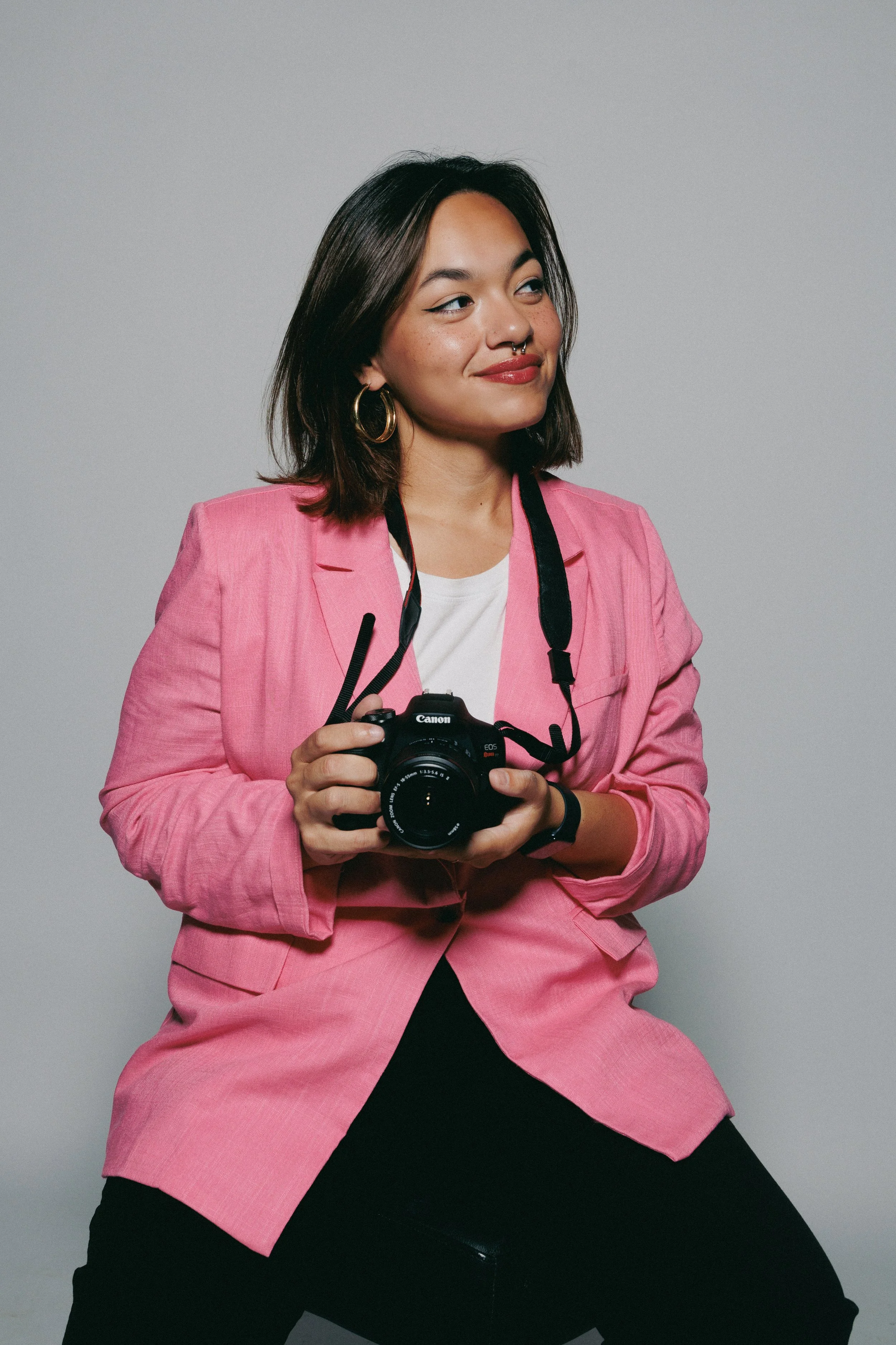 A woman in a pink blazer and black pants sitting on a black stool, holding a Canon camera, with a slight smile and looking to the side against a plain gray background.