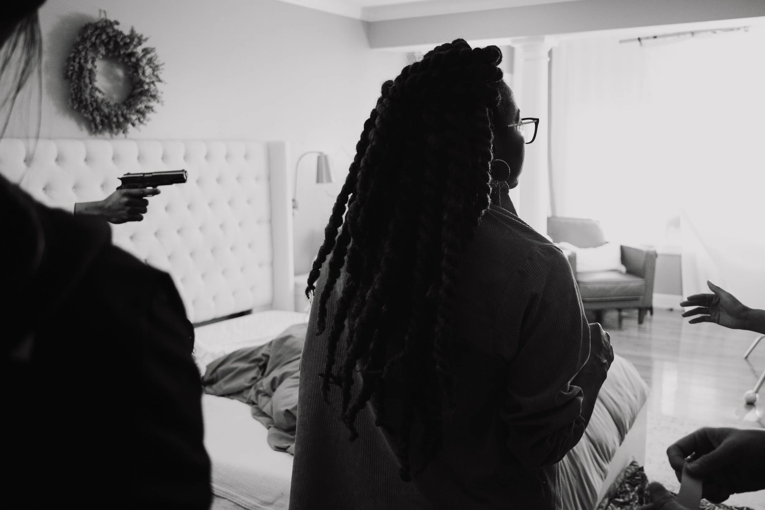 In this black-and-white image, a woman with long, textured hair, glasses, and hoop earrings stands in profile. She is surrounded by other people, one of whom is pointing a gun at her in a threatening manner. The scene appears to be indoors, in a room