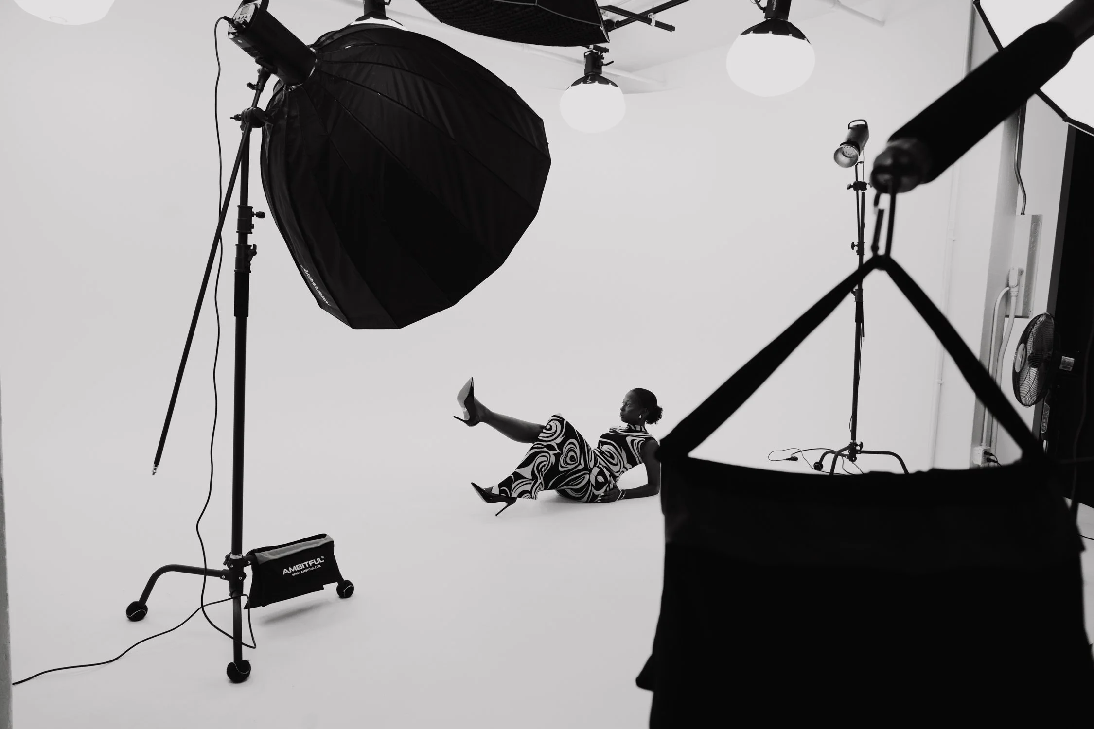 Fashion model posing on photography set with studio lighting and equipment in black and white.