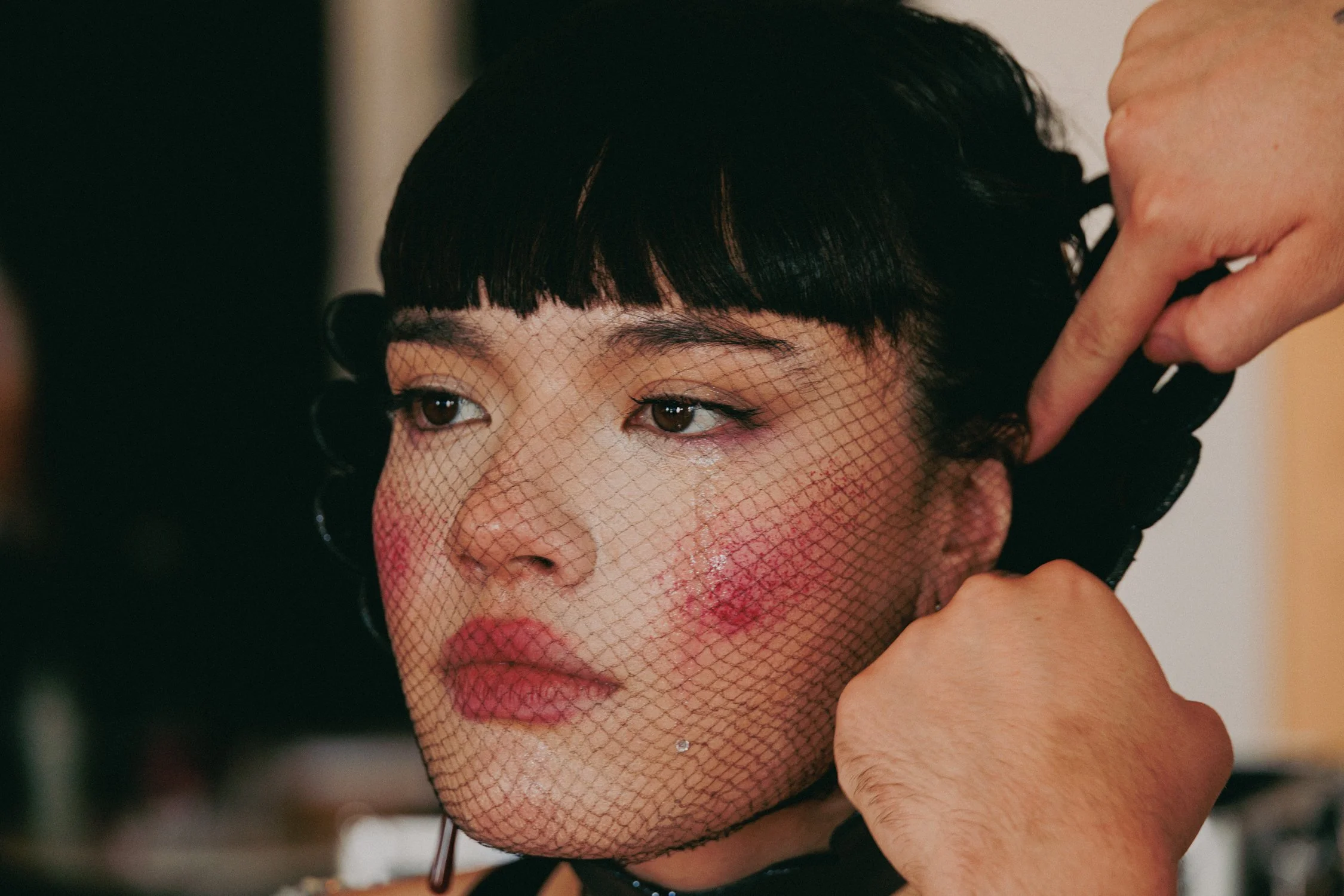 Close-up of a woman with short black hair, makeup, and a netted veil, as she adjusts her hair with her hand.