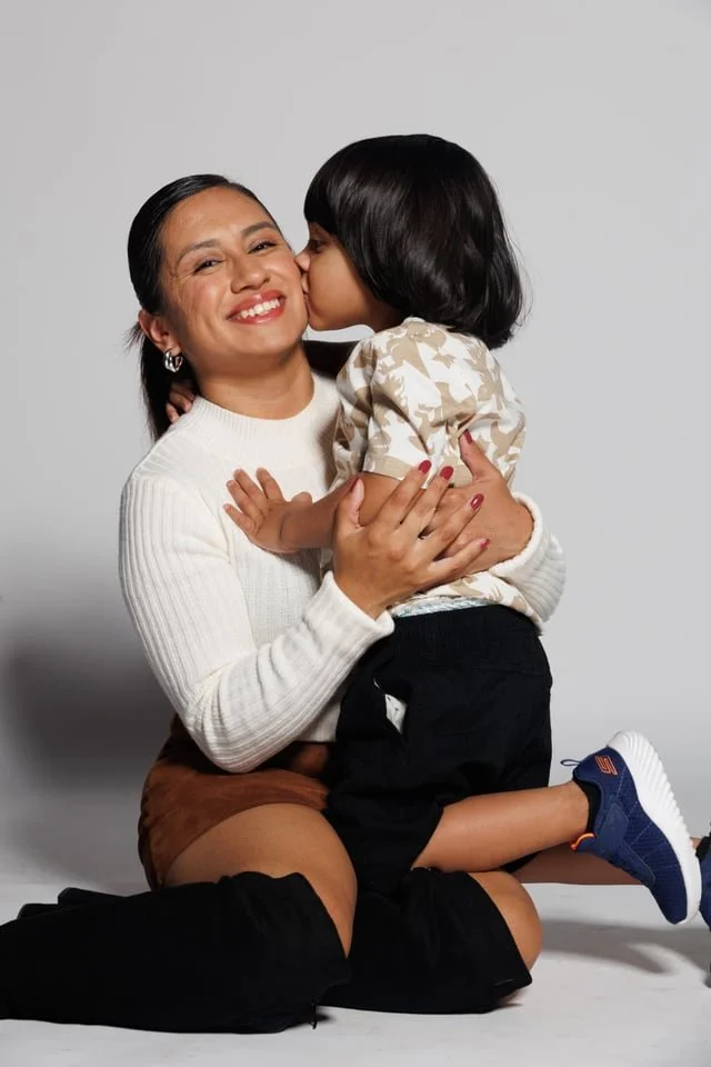 A woman with long dark hair smiling and holding a young girl, who is giving her a kiss on the cheek, against a plain gray background.