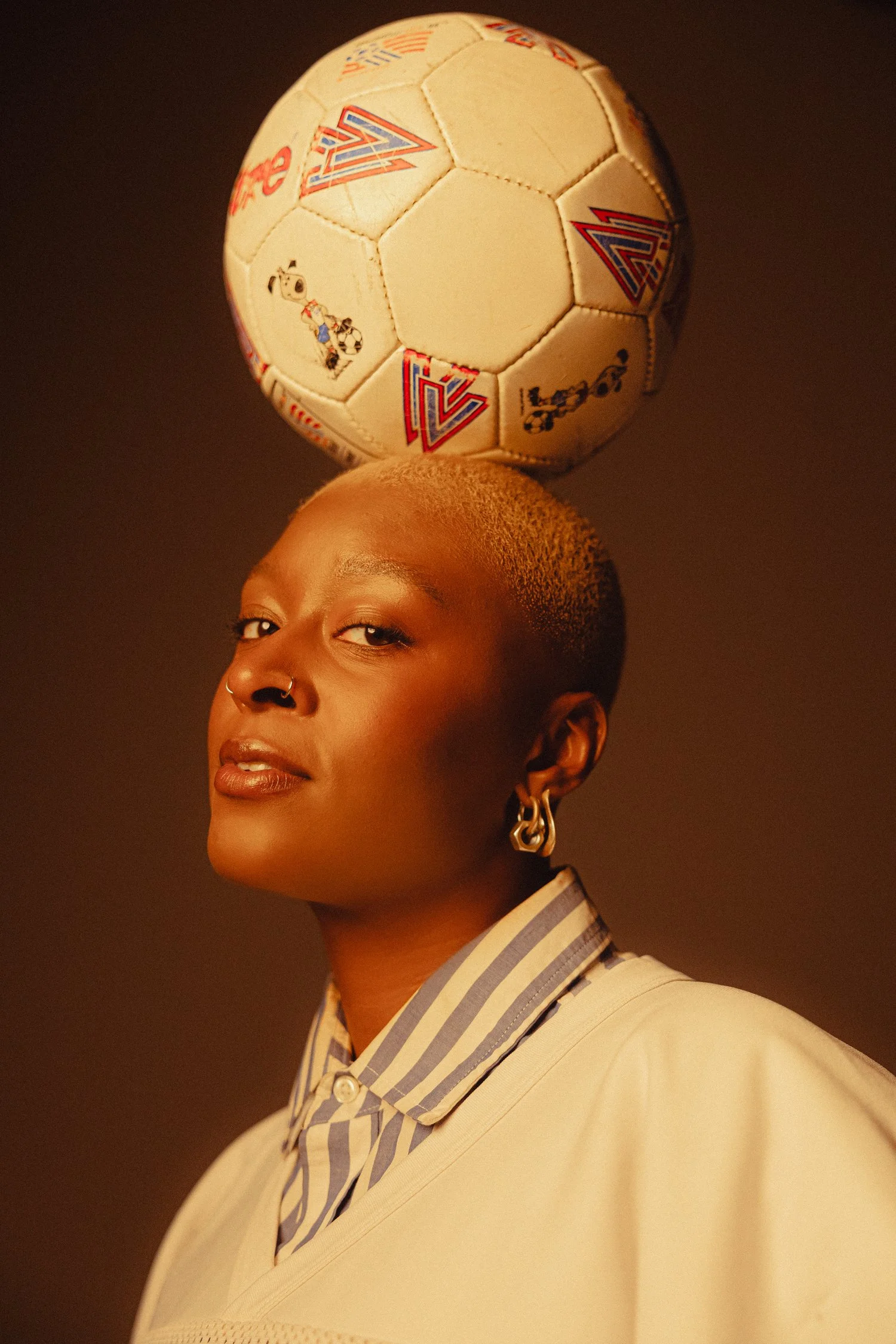 A young woman with short hair, wearing a striped shirt and jewelry, balancing a small soccer ball on her head against a dark background.