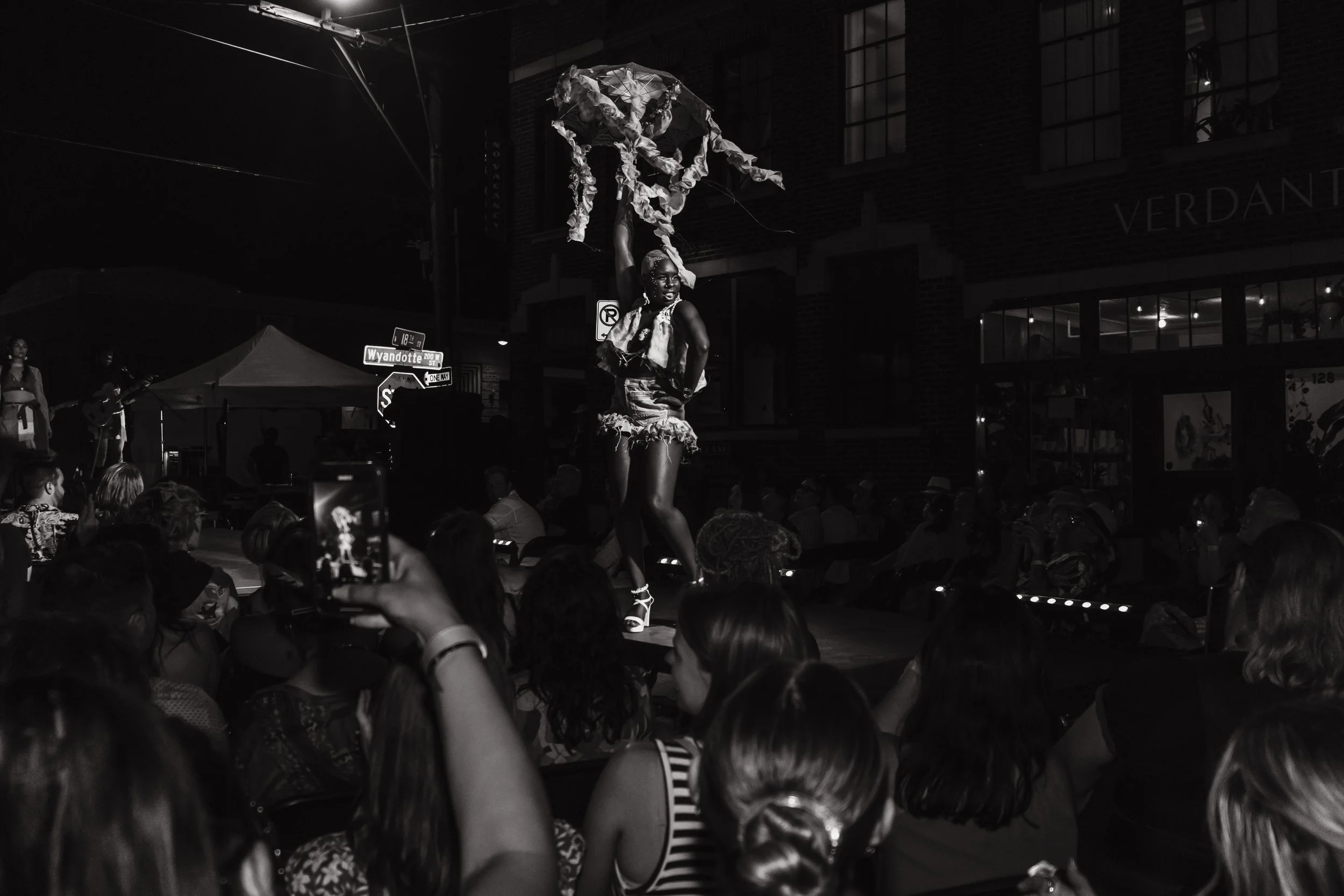 A performer dressed in decorative clothing is on stage, holding a large cloth above their head, with an audience watching. The scene is set outdoors at night, with some buildings and street signs visible in the background.