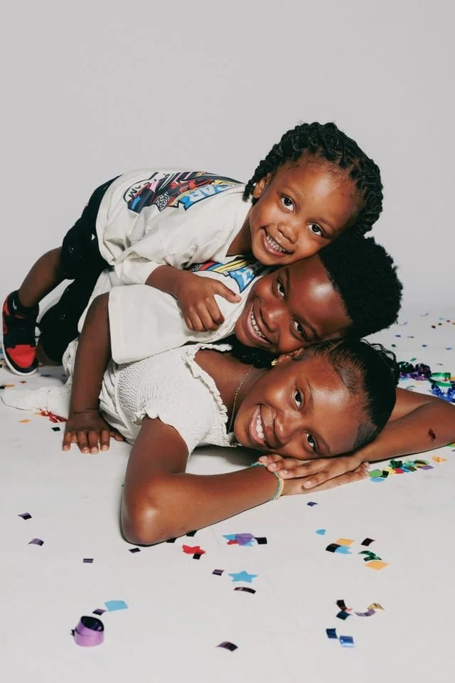 Three children smiling and playing on a white surface surrounded by colorful confetti, with a plain gray background.